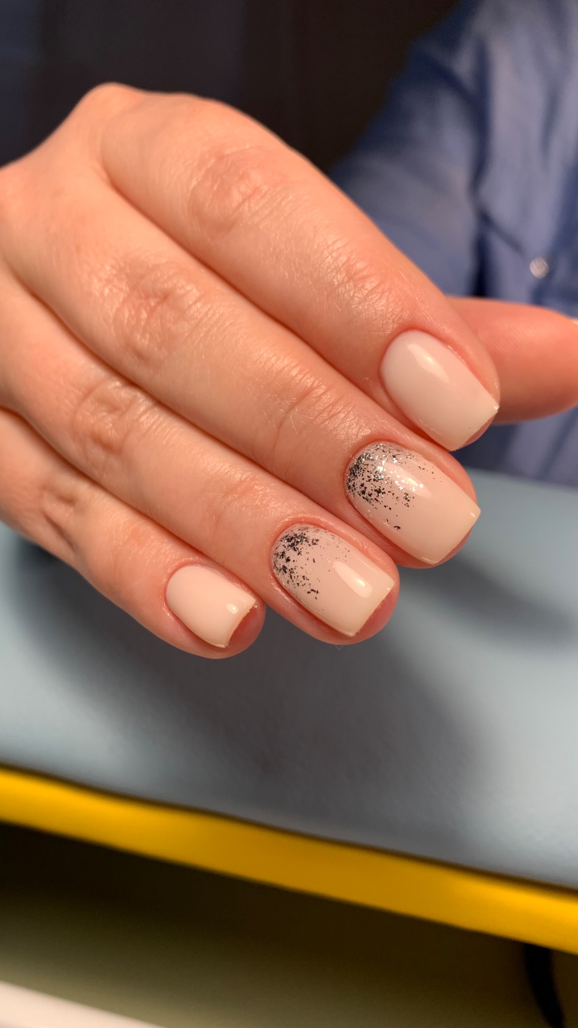 Dłoń z krótkimi, beżowymi paznokciami; dwa paznokcie zdobione srebrnym brokatem u nasady, na błękitnym tle.