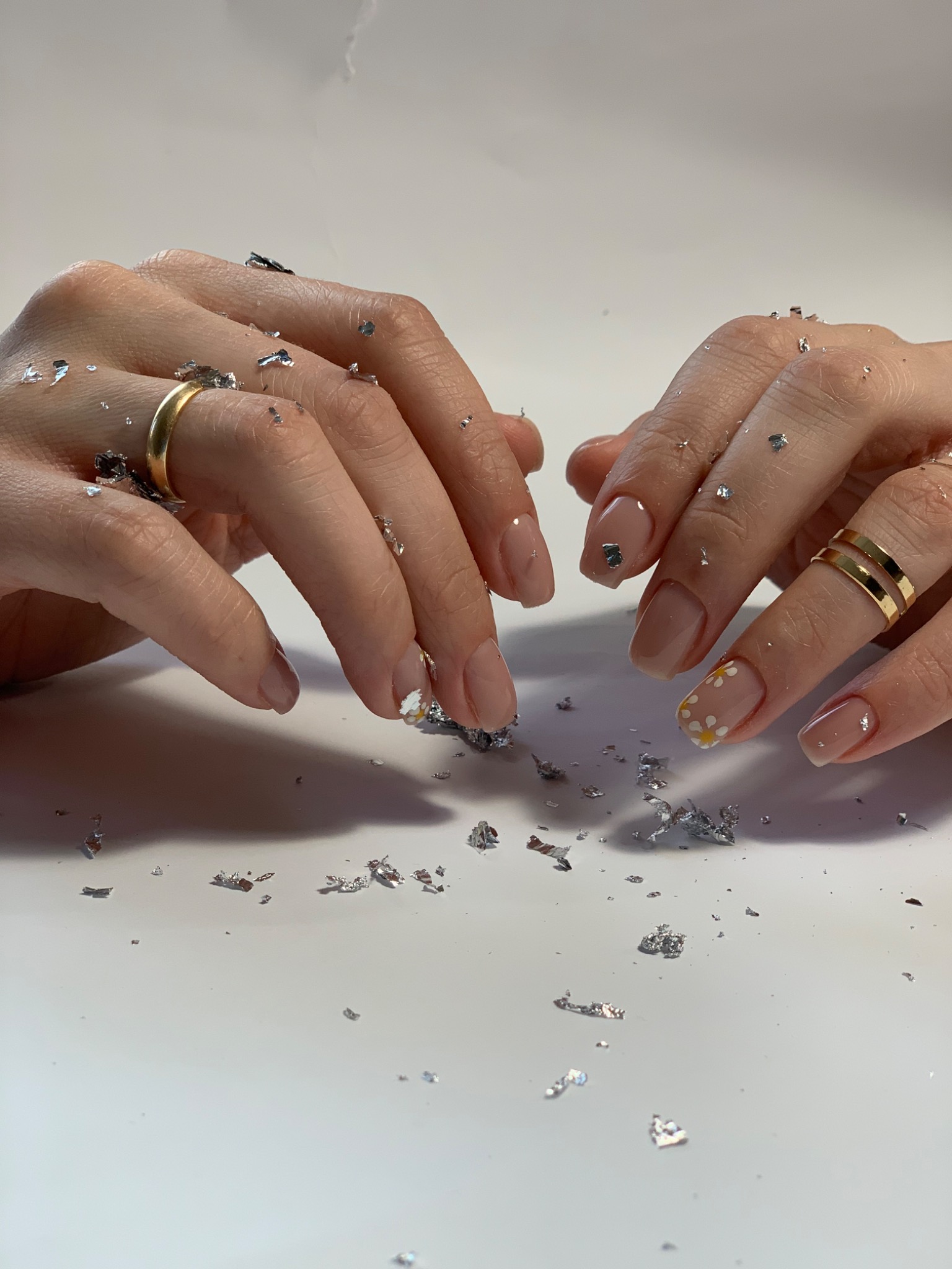 Dłonie z delikatnym, naturalnym manicure hybrydowym, jeden paznokieć ozdobiony drobnymi, białymi kwiatkami, otoczone srebrnymi płatkami folii.