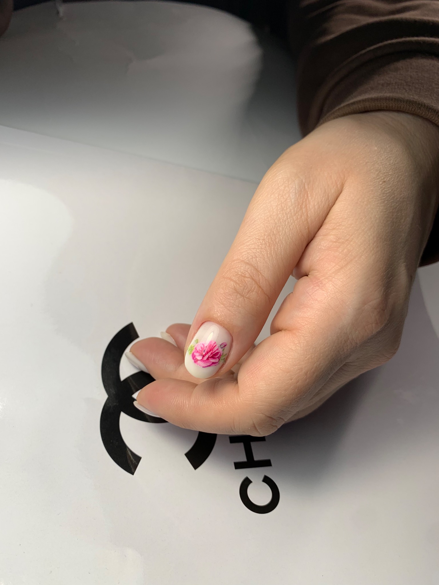 Powiększone ujęcie dłoni z jednym pomalowanym paznokciem w kolorze kremowym, zdobionym różową piwonią, na tle białego opakowania z czarnym logo Chanel.