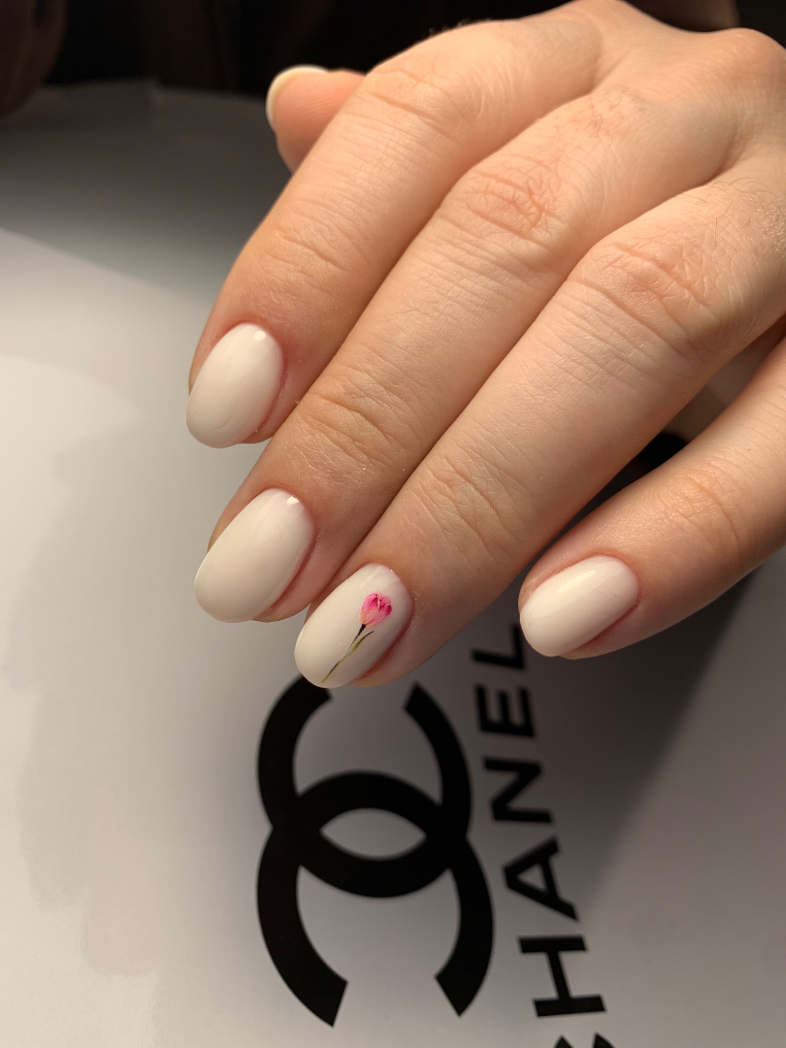 Kremowe paznokcie hybrydowe o owalnym kształcie, jeden z nich ozdobiony małym, różowym kwiatkiem, na tle z logo Chanel.