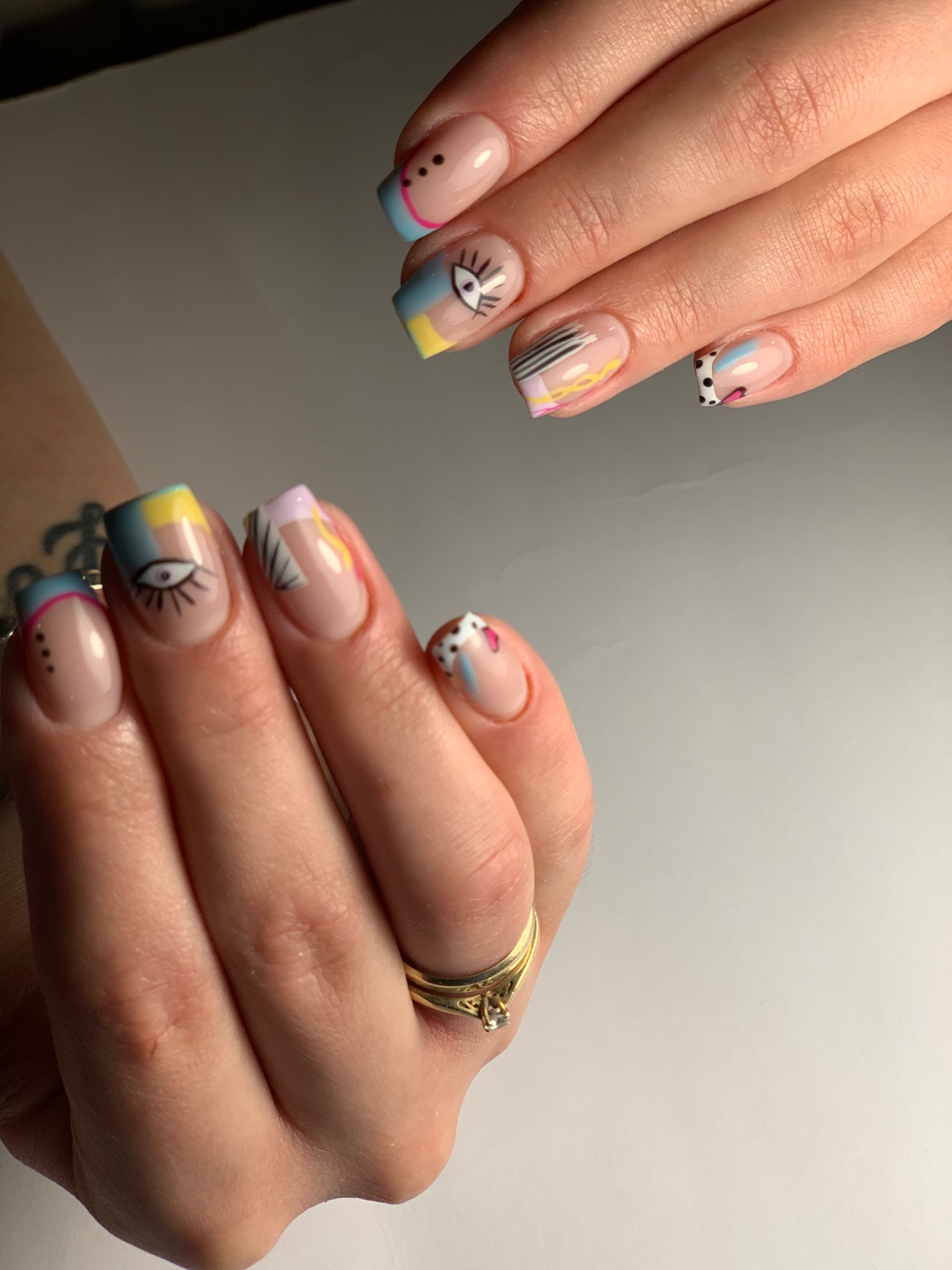 Dłonie z kwadratowymi paznokciami, na których widnieją abstrakcyjne, kolorowe wzory, w tym motyw oka, na tle białego stołu.