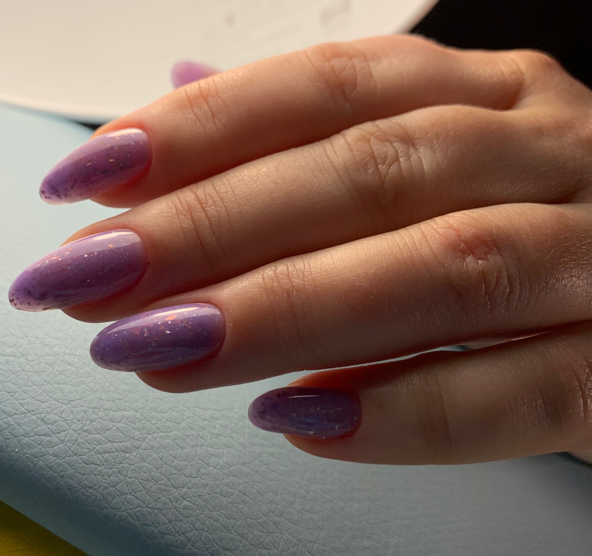 Dłoń z owalnymi paznokciami pomalowanymi na fioletowo-różowy kolor z efektem brokatu, ułożona na jasnoniebieskim podłożu.