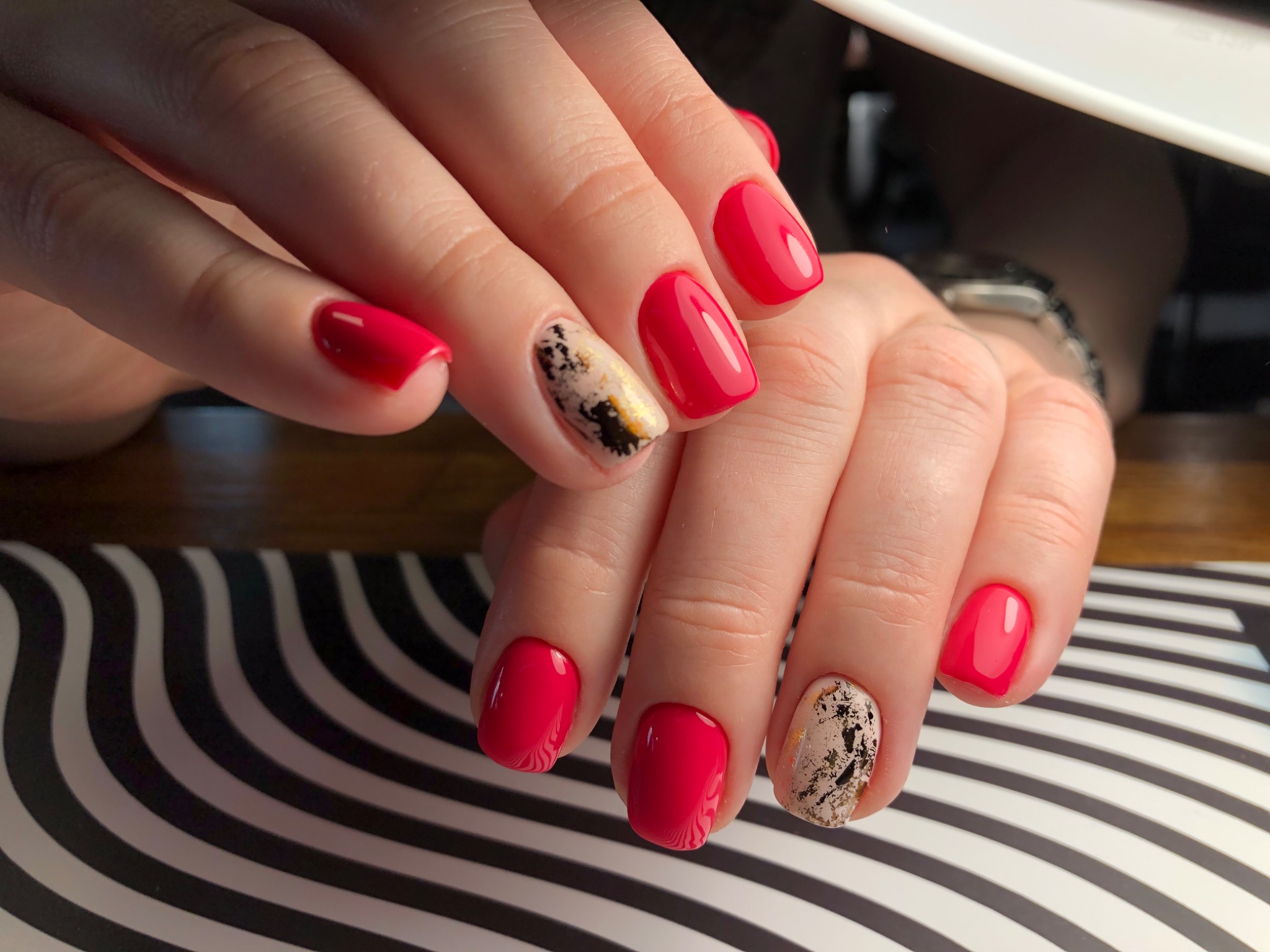 Dłonie z czerwonymi paznokciami i jednym paznokciem z abstrakcyjnym, złotym zdobieniem na tle czarno-białego wzoru.
