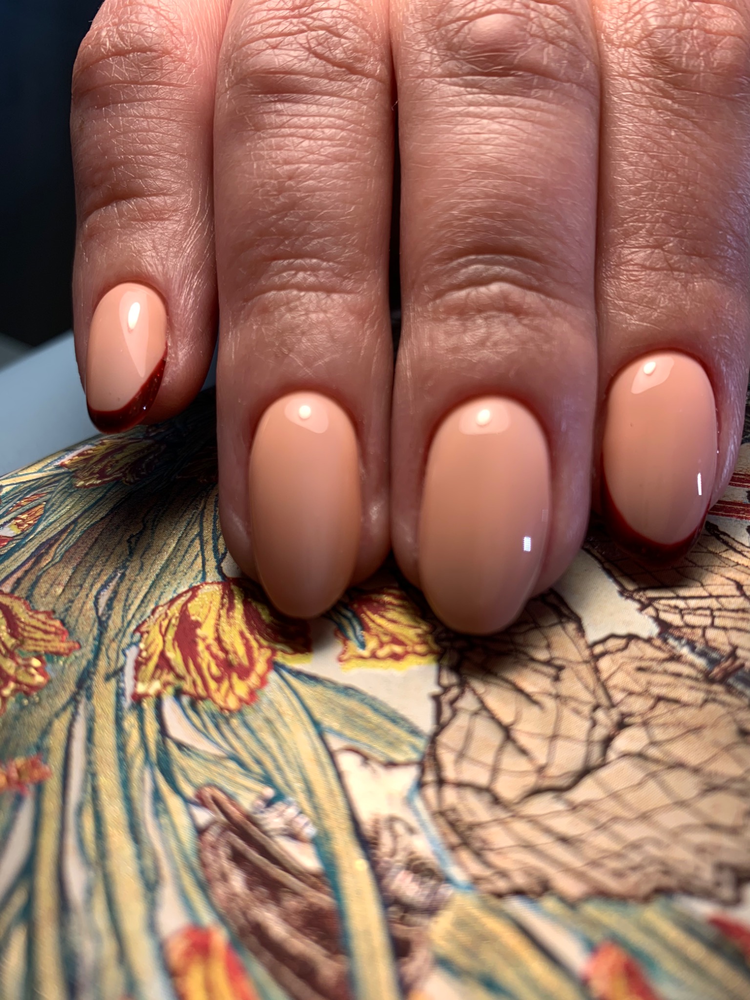 Zbliżenie dłoni z owalnymi paznokciami w odcieniu nude, jeden paznokieć z ciemnoczerwonym akcentem przy skórkach, na tle wzorzystej tkaniny.