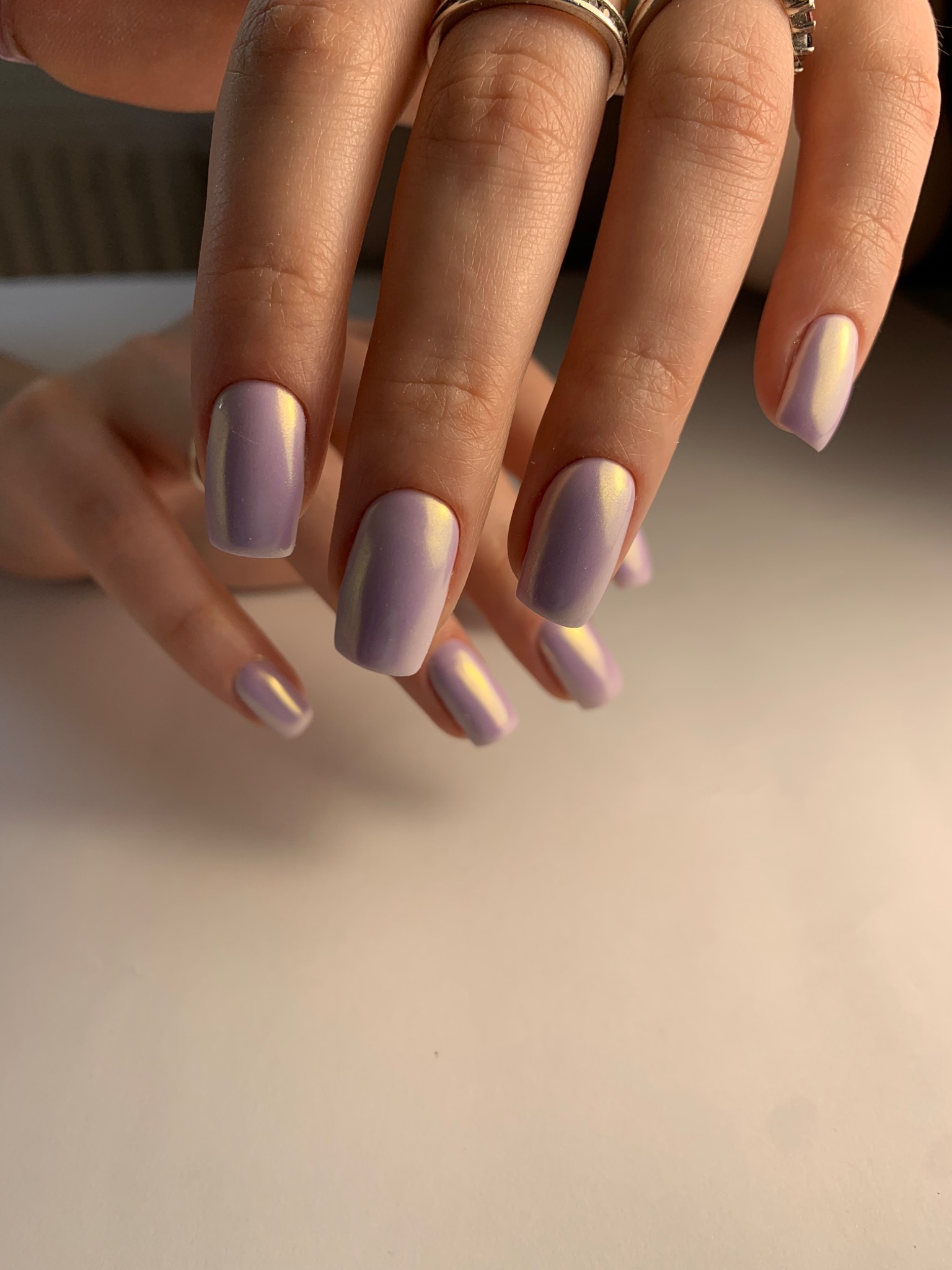 Delikatne, liliowe paznokcie z opalizującym wykończeniem, wykonane w salonie w Warszawie. Widoczne pierścionki na palcach.