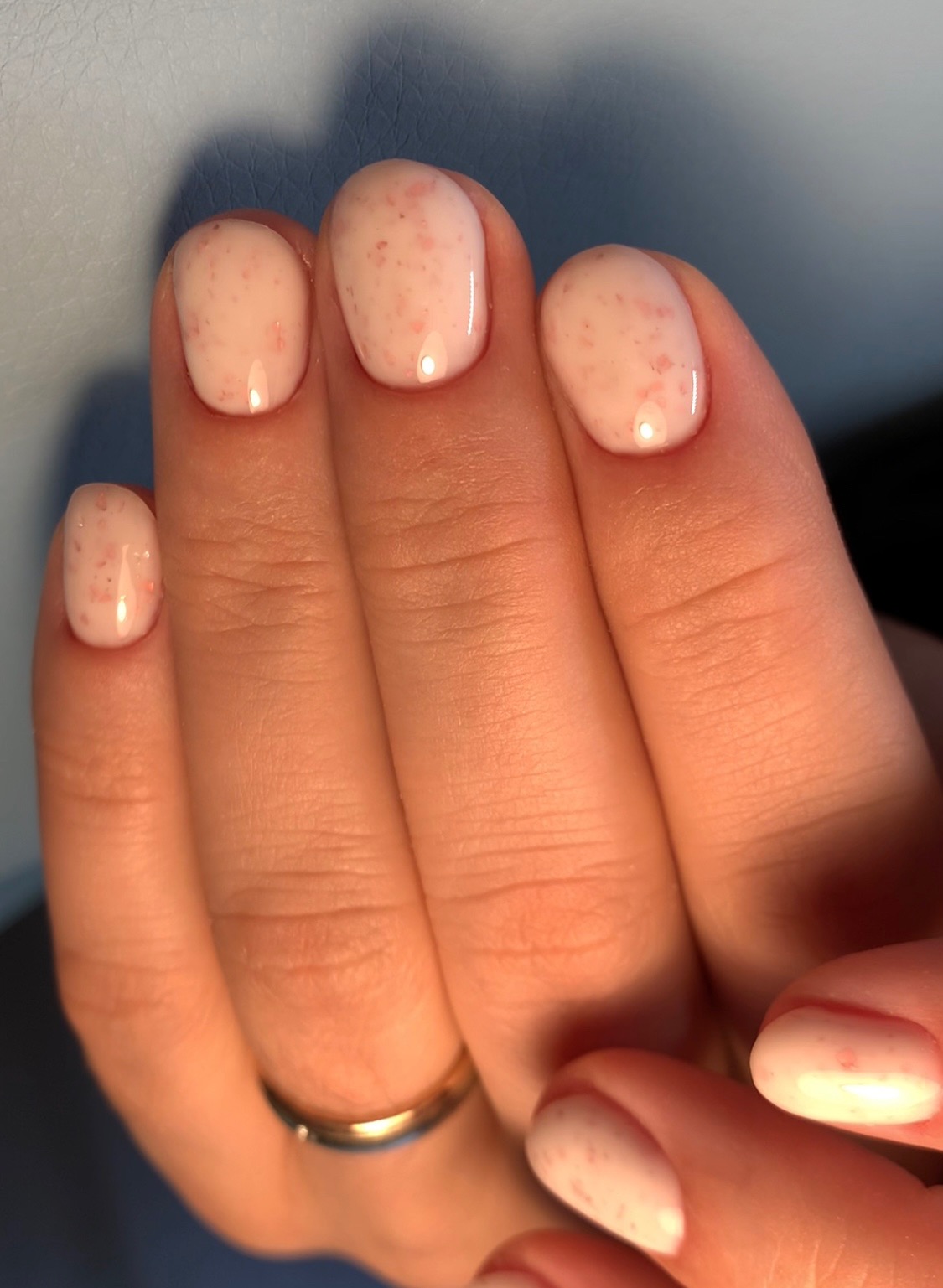 Dłoń z owalnymi paznokciami pomalowanymi jasnoróżowym lakierem z drobnymi, nieregularnymi plamkami w kolorze różowym, na serdecznym palcu złoty pierścionek.