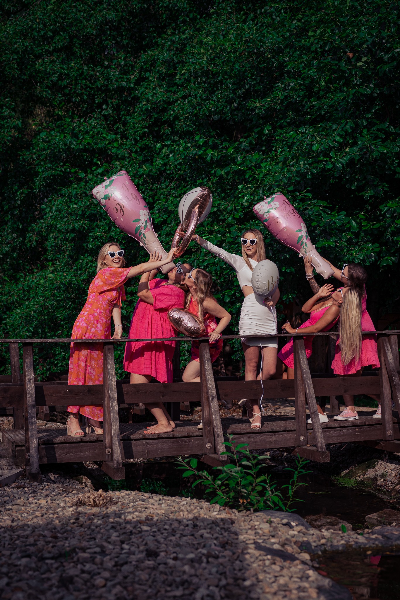 Grupa kobiet w różowych sukienkach i białej sukience na drewnianym moście, trzymające balony w kształcie butelek szampana i pierścieni, świętująca wieczór panieński na tle zieleni.