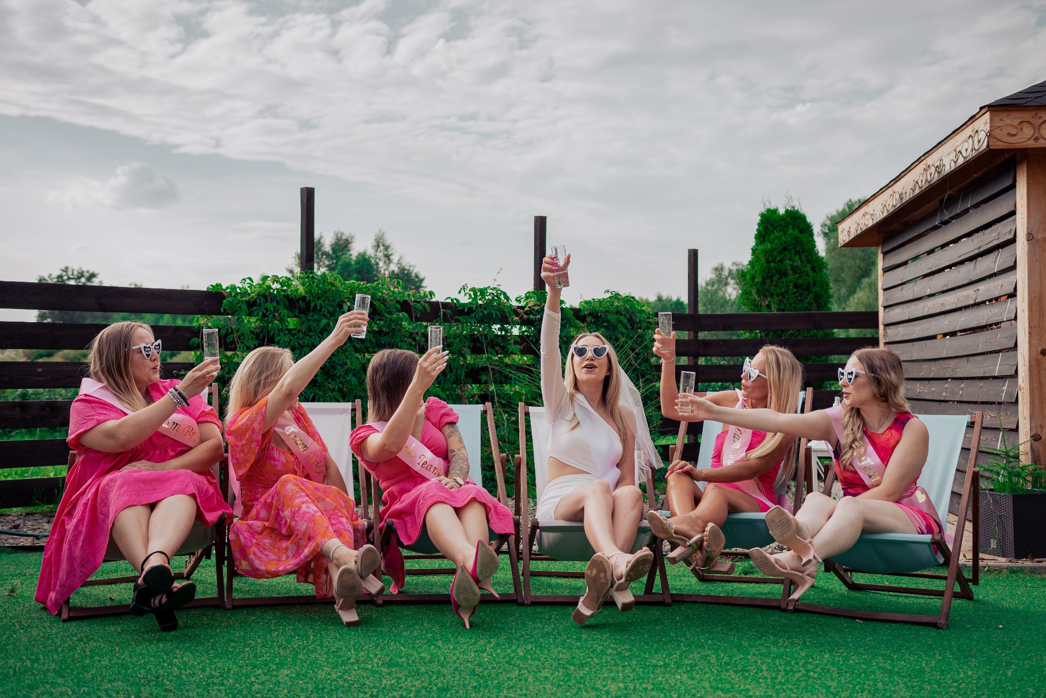 Grupa kobiet w różowych sukienkach i szarfach 'Team Bride' siedzi na leżakach, wznosząc toasty szampanem podczas wieczoru panieńskiego na tle zielonego trawnika i drewnianej altanki.