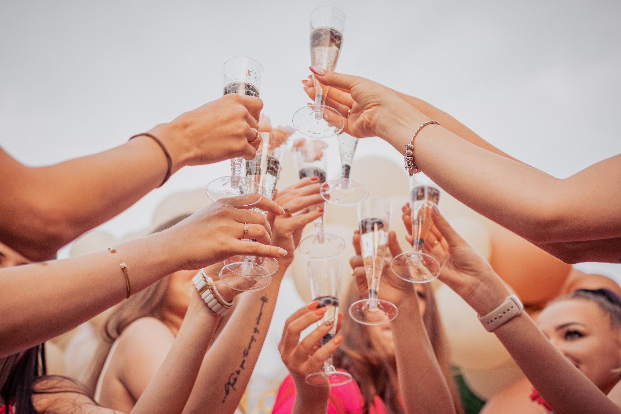 Grupa kobiet wznosi toast szampanem, widoczne dłonie z biżuterią i tatuażem, tło z balonami.