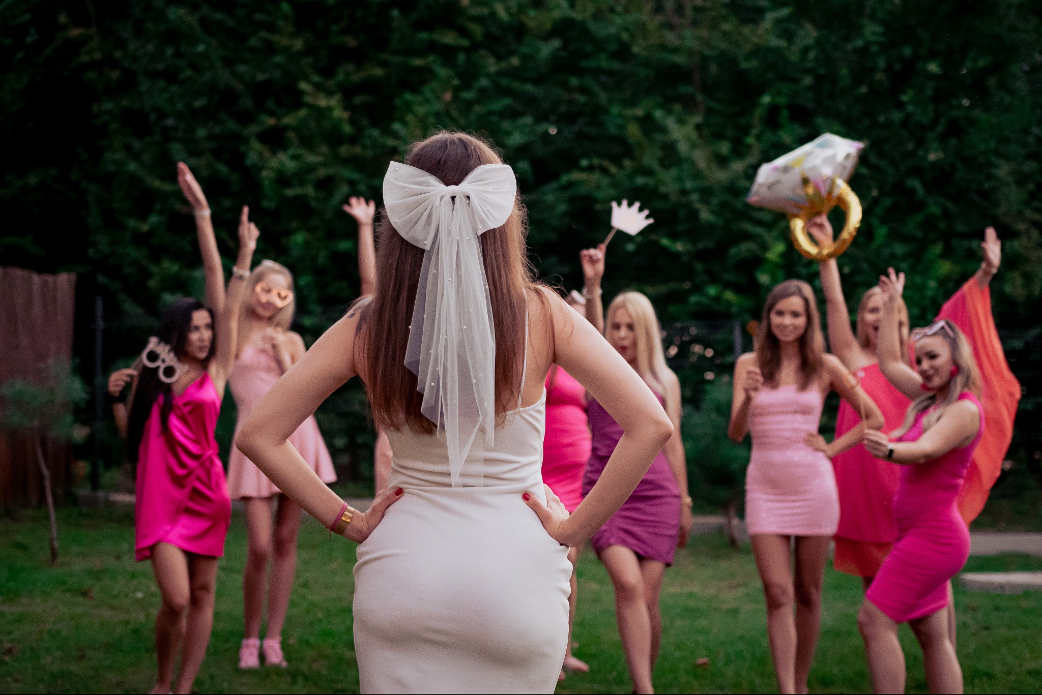 Panna młoda tyłem, z kokardą we włosach i welonem z perełkami, otoczona przez rozbawione druhny w różowych sukienkach z rekwizytami, takimi jak dmuchany pierścionek i korona, na tle zieleni.