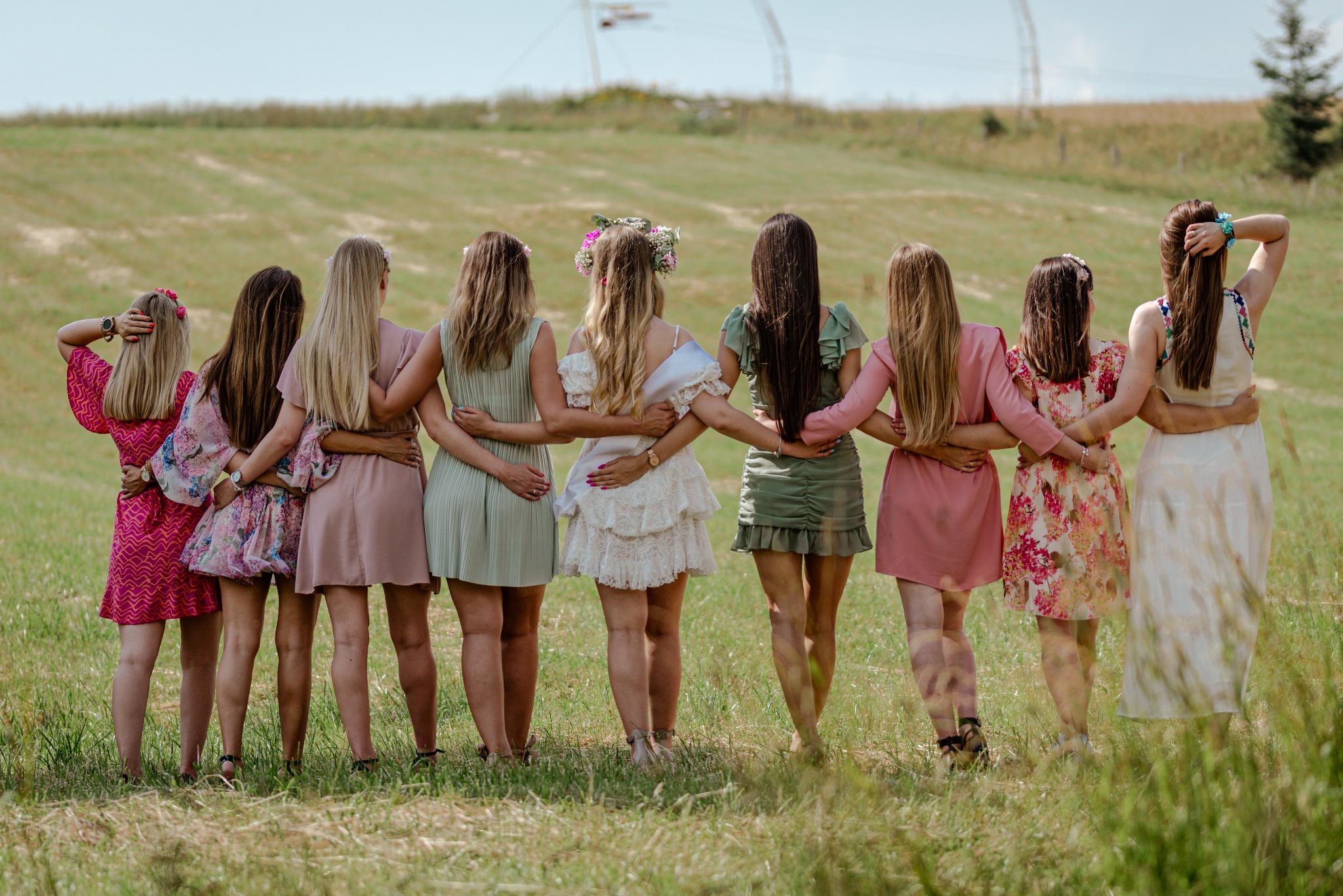 Grupa kobiet, tyłem, obejmujących się na tle zielonego pola, prawdopodobnie uczestniczki wieczoru panieńskiego, z jedną z nich w białej sukience i wianku.