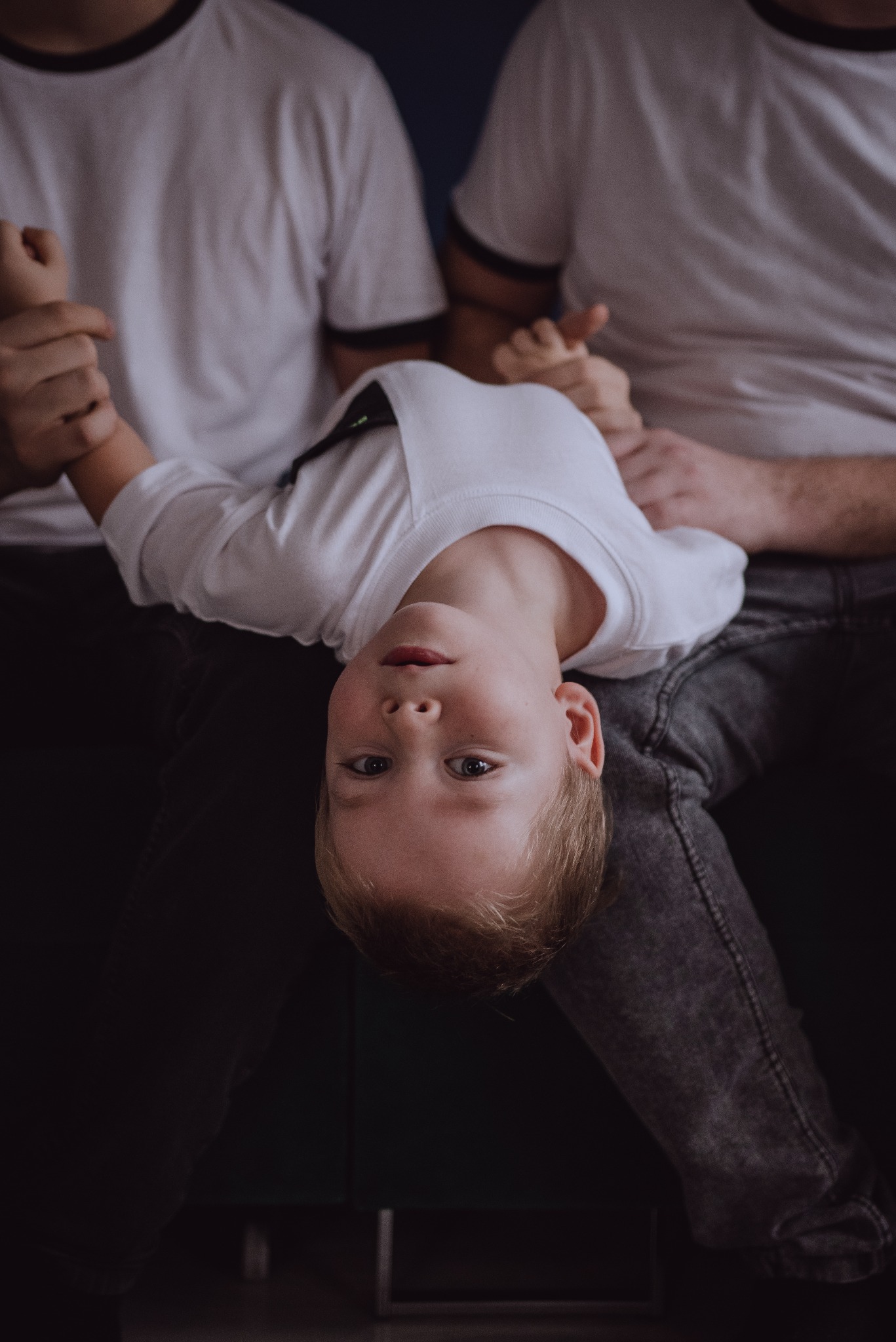 Chłopiec w białej koszulce, trzymany przez dwóch mężczyzn, zwisa głową w dół, patrząc w obiektyw.