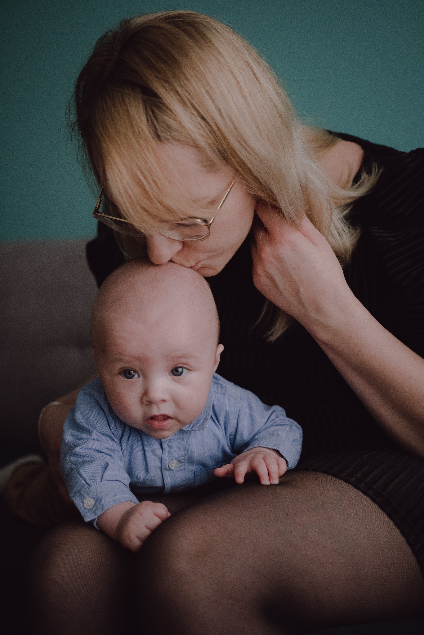 Matka w okularach całuje w głowę niemowlę ubrane w błękitną koszulę na tle turkusowej ściany.