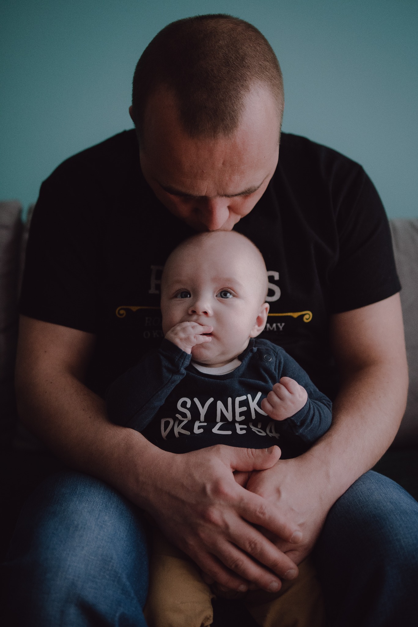 Ojciec całuje w głowę niemowlę trzymane na kolanach, dziecko w granatowym body z napisem 'Synek Prezesa' patrzy w obiektyw, tło stanowi turkusowa ściana.
