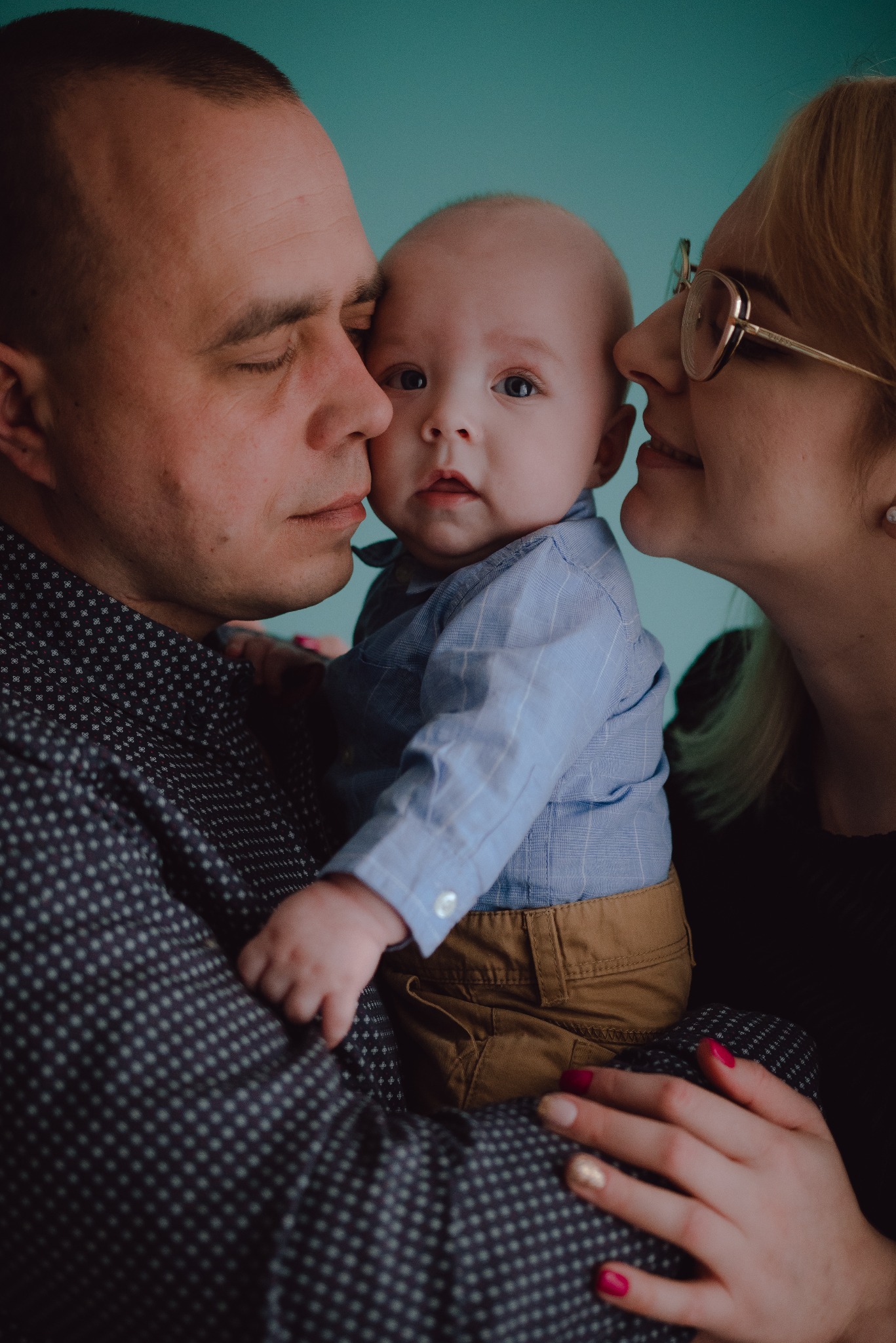 Rodzice tulący niemowlę w studio na tle turkusowej ściany, matka w okularach, dziecko w błękitnej koszuli, delikatne oświetlenie.