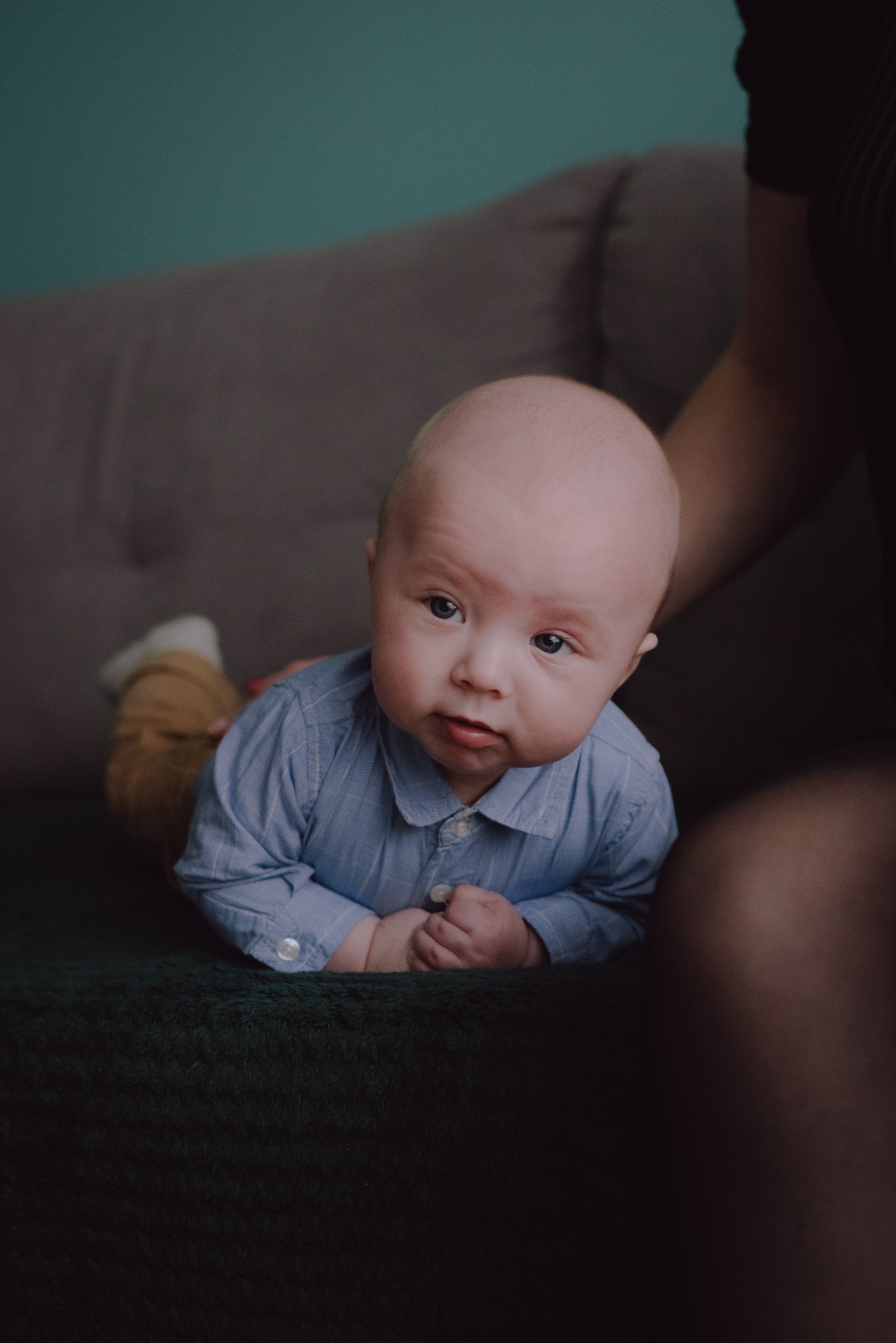 Niemowlę w błękitnej koszuli leży na brzuchu na ciemnozielonym kocu, wsparte na dłoniach, z poważnym wyrazem twarzy, w tle fragment sofy i ściany.