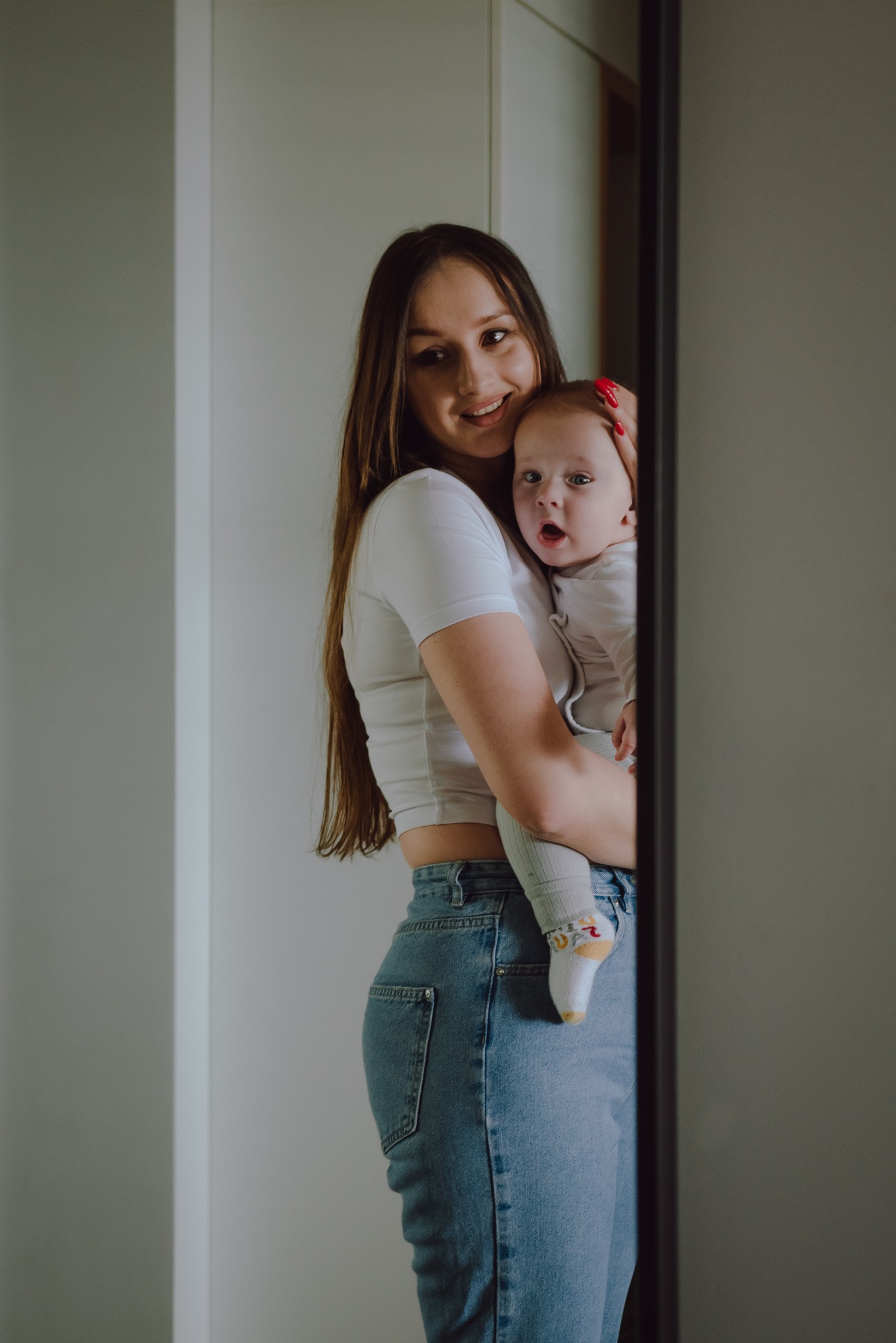 Uśmiechnięta młoda mama w białym crop topie i jeansach trzyma na rękach niemowlę w białym ubranku i skarpetkach z kolorowymi wzorami, stojąc na tle białej ściany i lustra.