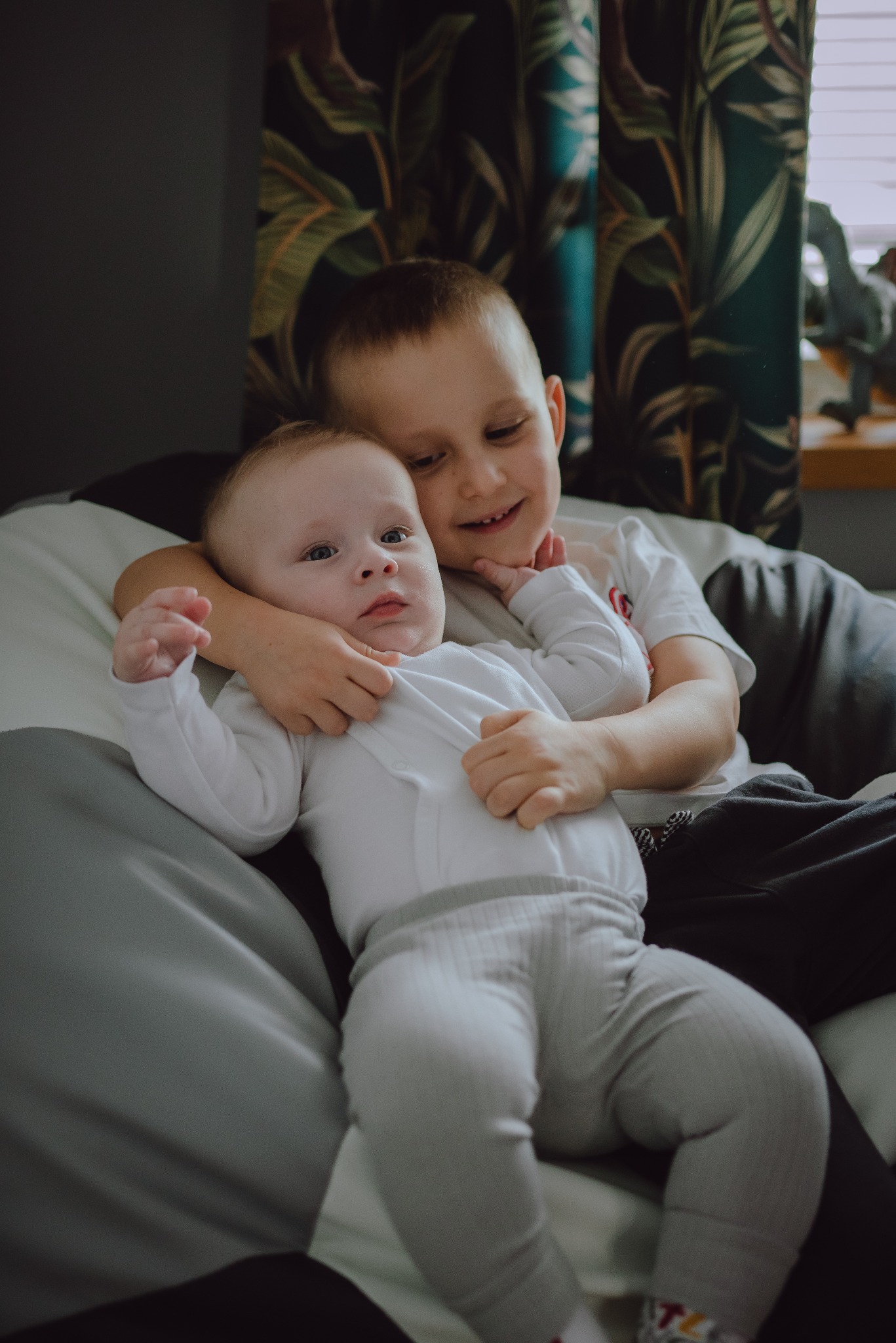 Uśmiechnięty starszy brat obejmuje młodszego, leżącego na poduszce w pokoju z zasłonami w tropikalne liście i figurką dinozaura na parapecie.