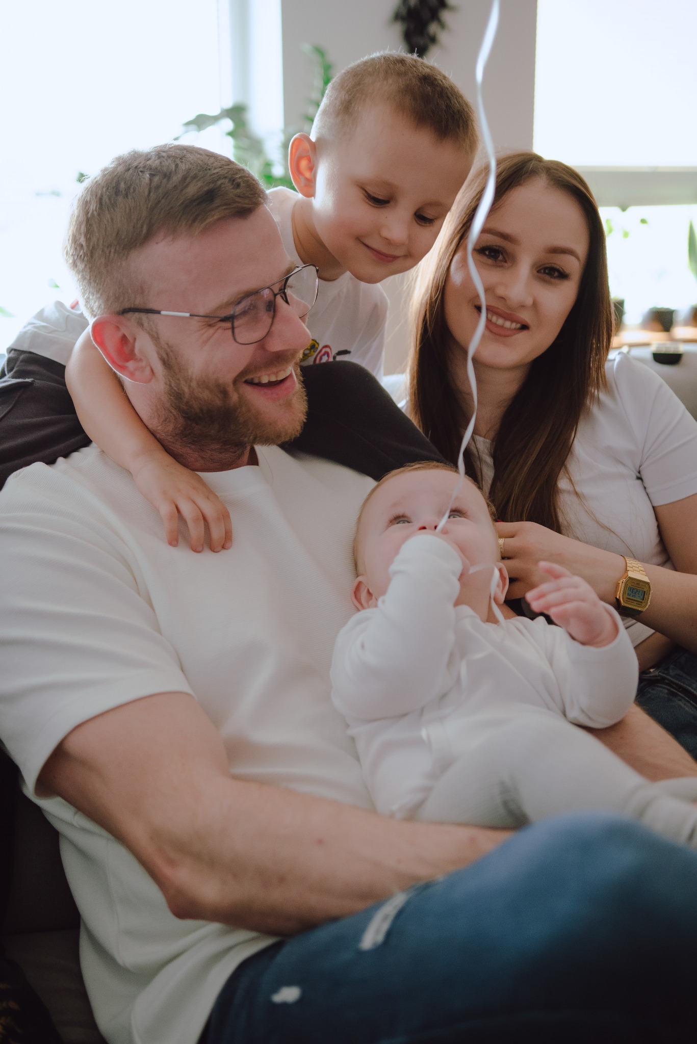 Uśmiechnięta rodzina: tata w okularach trzyma niemowlę w białym ubranku, chłopiec opiera się na jego ramionach, a mama z zegarkiem na ręce uśmiecha się w tle, dziecko bawi się sznurkiem od balona.