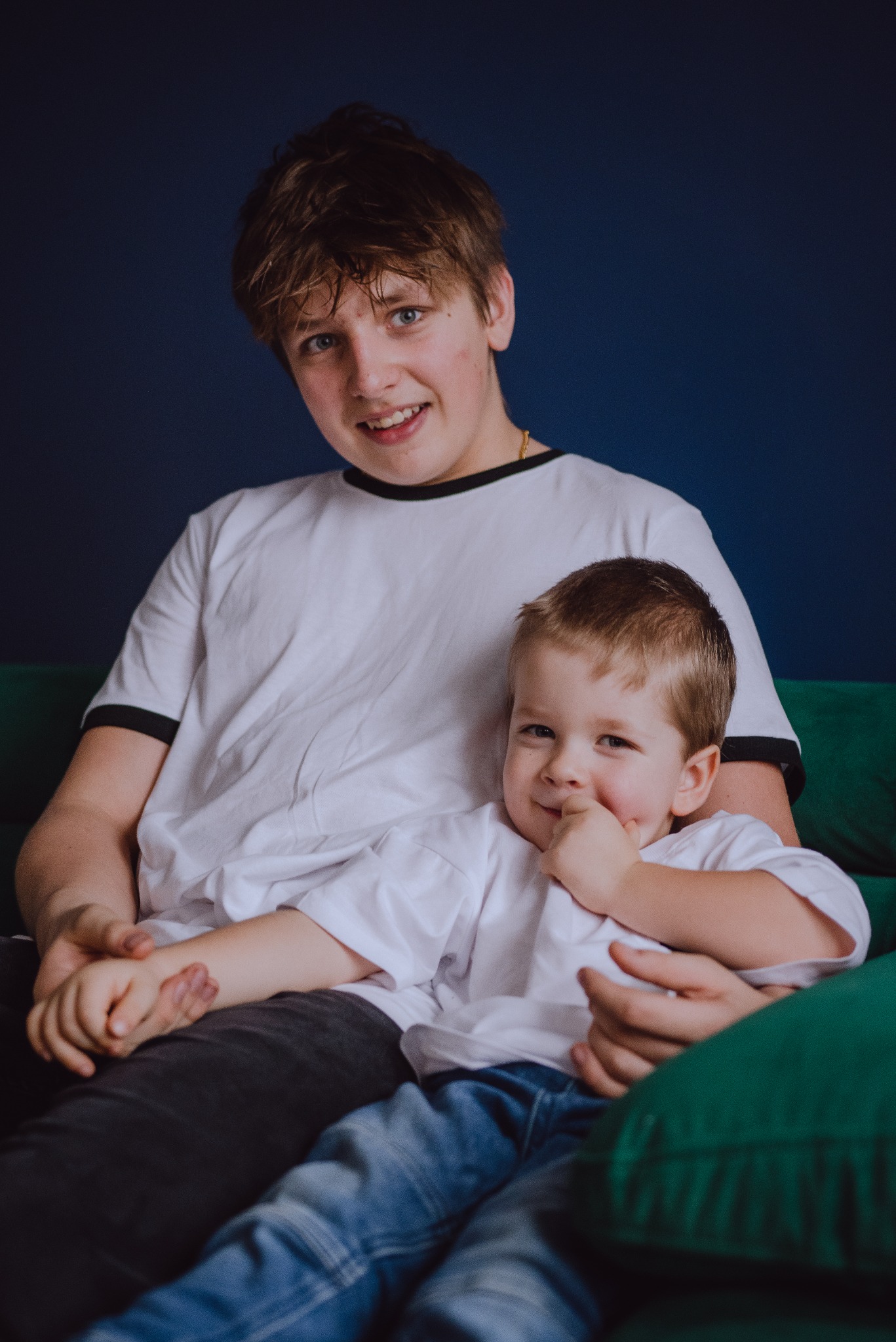 Portret dwóch chłopców: starszy nastolatek obejmuje młodszego brata, siedząc na zielonej sofie na tle ciemnoniebieskiej ściany. Młodszy chłopiec uśmiecha się, trzymając palec przy ustach.