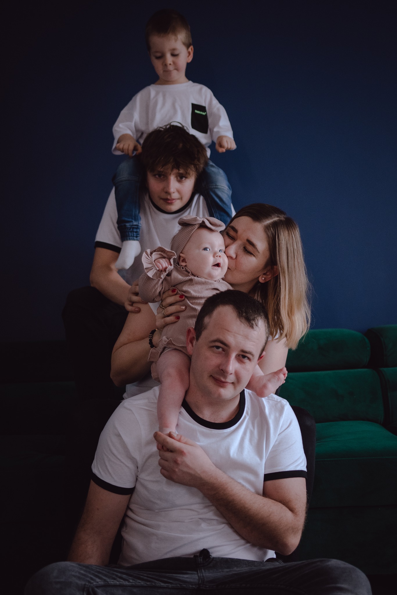 Uśmiechnięta rodzina: tata z córeczką na ramionach, mama całuje córeczkę, za nimi nastoletni syn z młodszym bratem na barana, wszyscy na tle ciemnoniebieskiej ściany i zielonej kanapy.