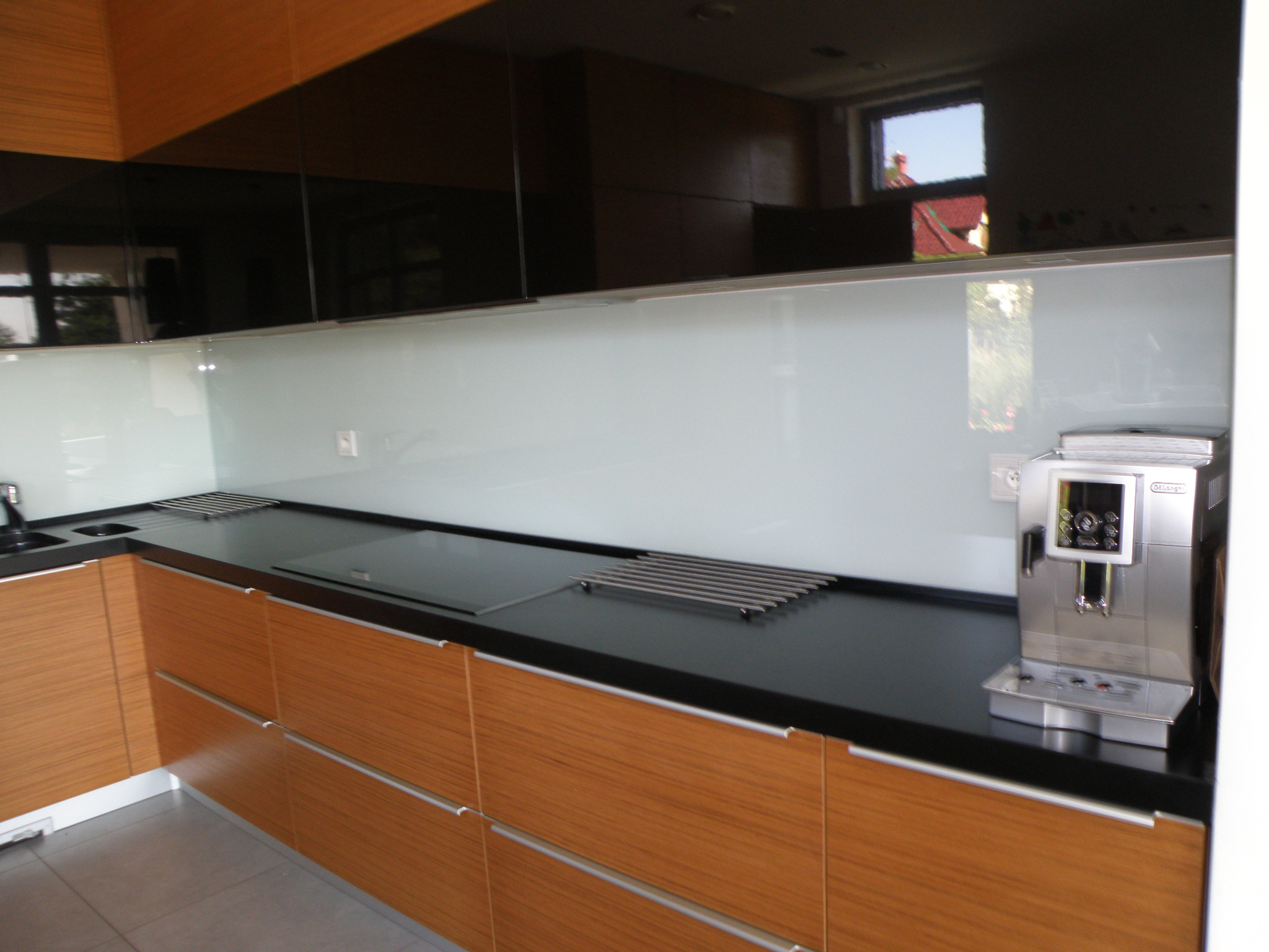 Biała szklana ściana Lacobel w kuchni, zamontowana nad blatem z zabudowaną płytą grzewczą i metalowym ociekaczem, z widocznym ekspresem do kawy.