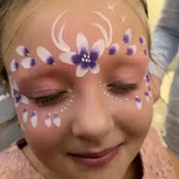 Dziewczynka z zamkniętymi oczami i pomalowaną twarzą w kwiatowe wzory w odcieniach bieli i fioletu z brokatowymi akcentami.