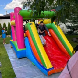 Para w strojach ślubnych zjeżdża z kolorowej, dmuchanej zjeżdżalni na trawniku, widoczne drzewa i goście w tle.