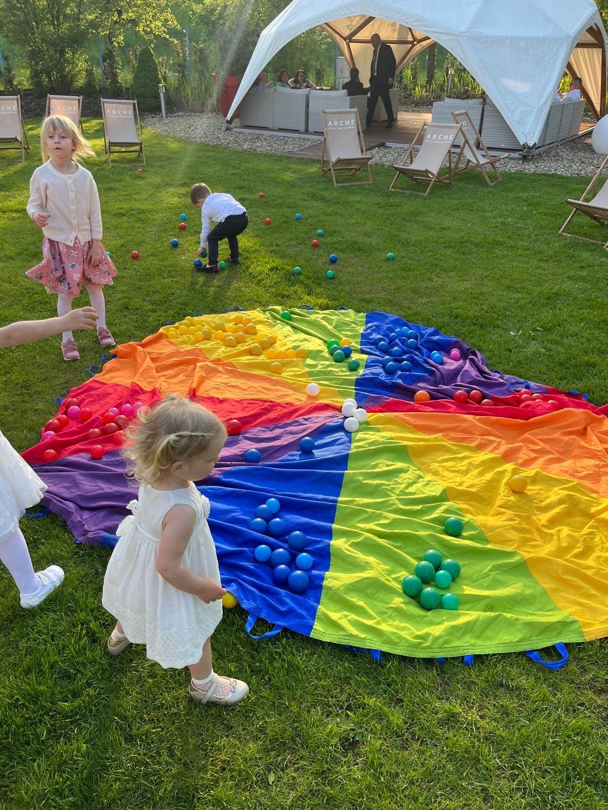 Dzieci bawią się na trawie kolorową chustą animacyjną wypełnioną plastikowymi piłeczkami; w tle biały namiot i krzesła.