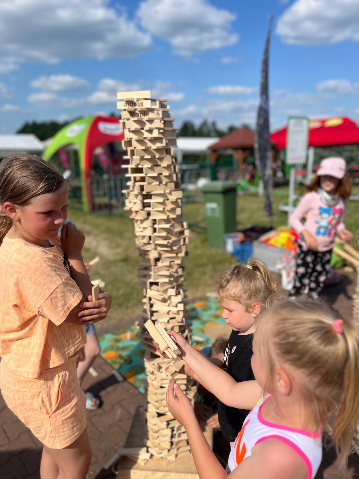 Dzieci budują wysoką wieżę z drewnianych klocków na zewnątrz, w tle widoczne namioty i inne atrakcje.