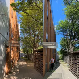 Mężczyzna w kamizelce odblaskowej sprząta po wycince gałęzi wzdłuż budynku z brązową elewacją, widoczne drzewa i niebo.