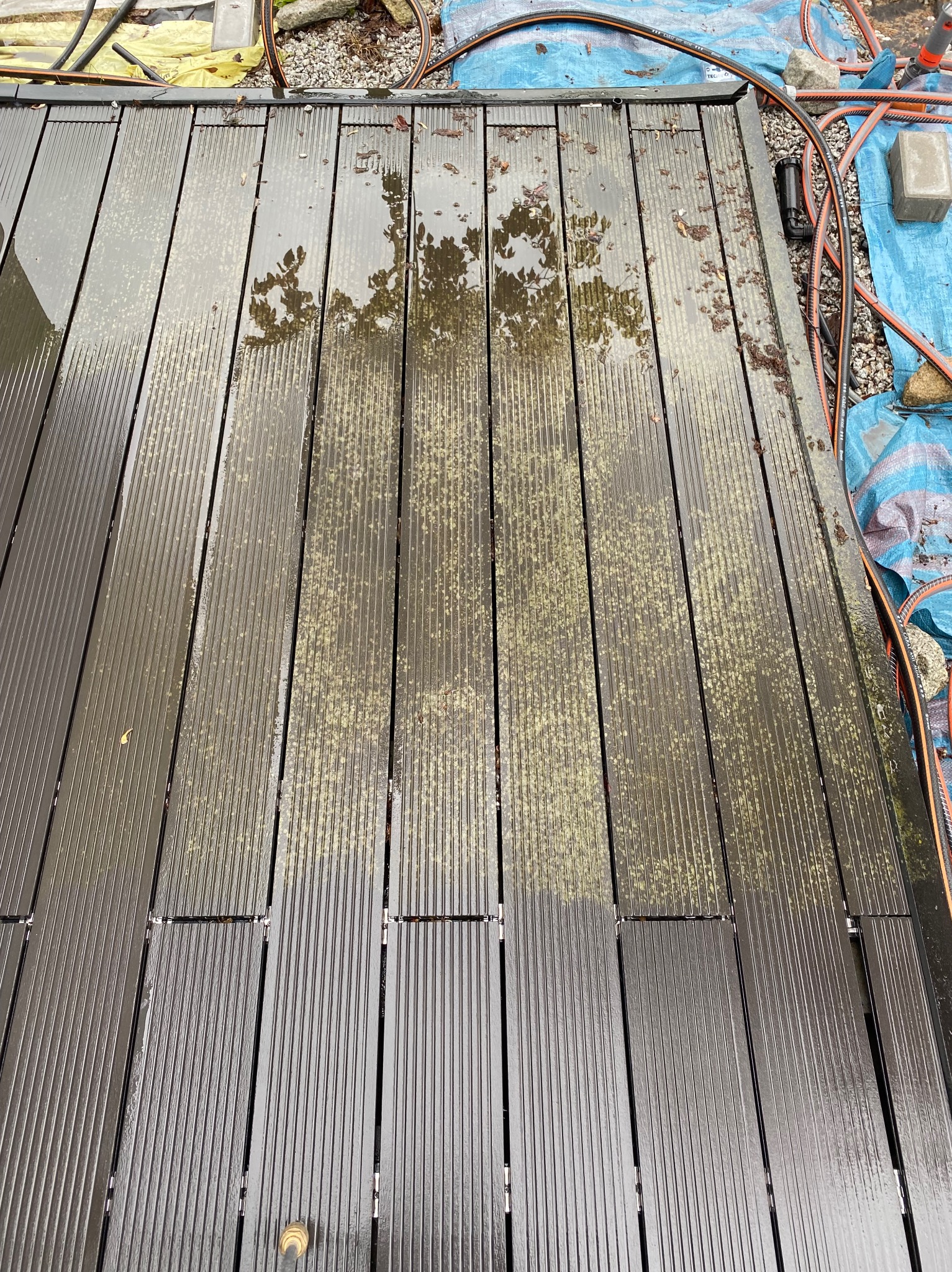 Częściowo umyta, mokra, brązowa deska tarasowa w Czerlejnku, widoczne pozostałości zielonego nalotu i liści, obok rozłożona niebieska plandeka i wąż ogrodowy.