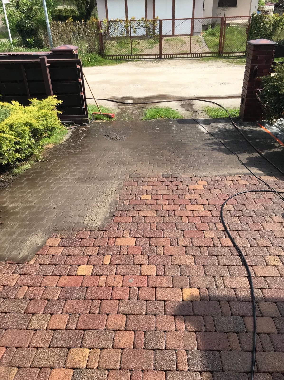 Częściowo umyta, czerwono-brązowa kostka brukowa na podjeździe, z widocznym brudem i wodą, obok brama wjazdowa i zieleń.