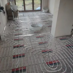 Rozłożone rury ogrzewania podłogowego na folii z siatką, widoczne zwoje rur, okno balkonowe i kartony w tle.