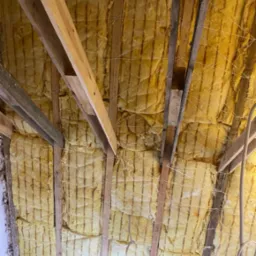 Żółta wełna mineralna zamontowana między drewnianymi krokwiami poddasza, zabezpieczona sznurkiem, widoczne fragmenty instalacji elektrycznej.