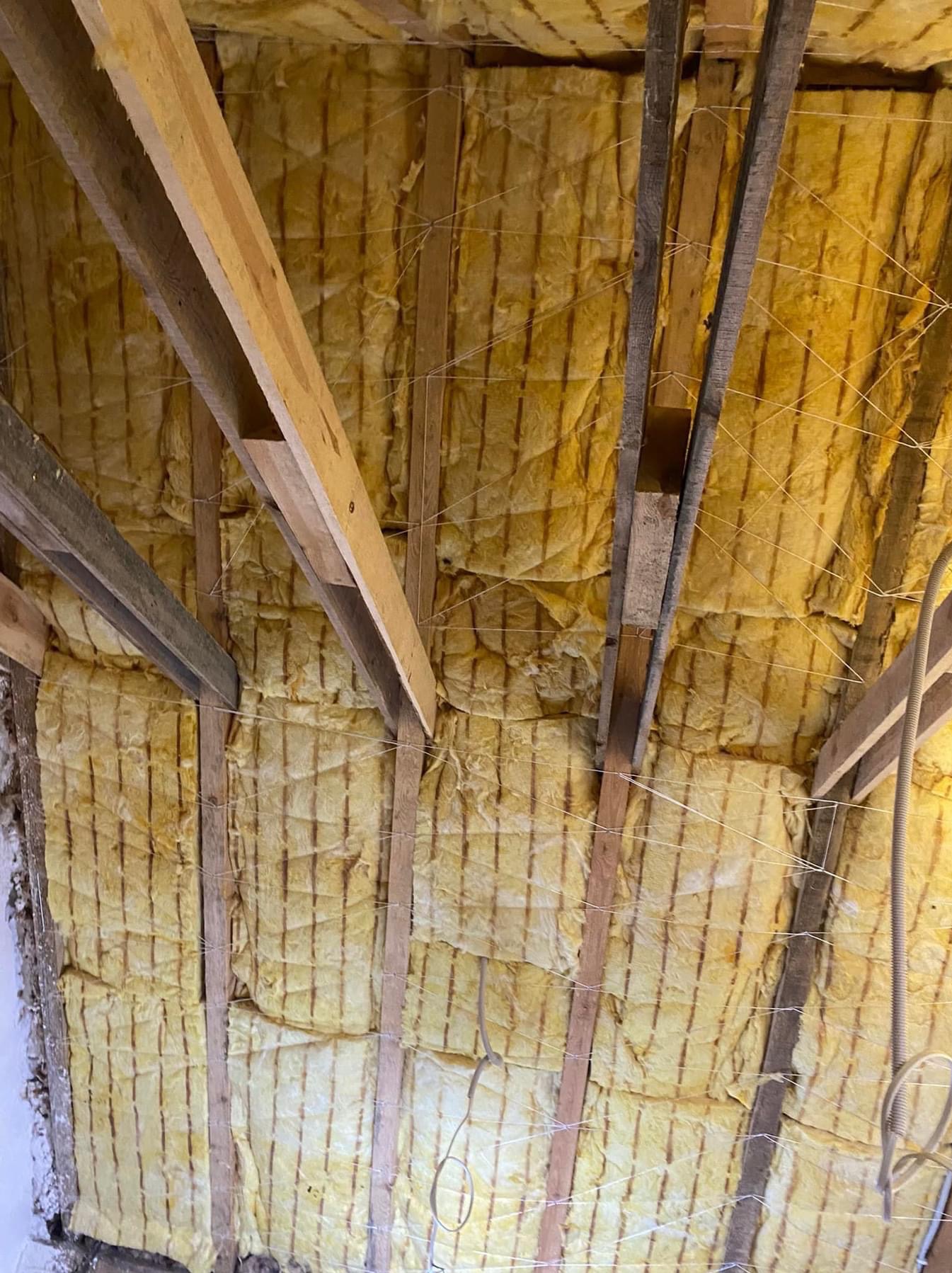 Żółta wełna mineralna zamontowana między drewnianymi krokwiami poddasza, zabezpieczona sznurkiem, widoczne fragmenty instalacji elektrycznej.
