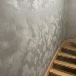 Ściana z efektem dekoracyjnym w odcieniach szarości i srebra, sąsiadująca z drewnianymi schodami o jasnym odcieniu, widoczne z boku.