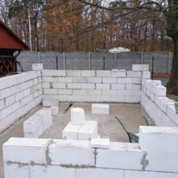 Wznoszenie ścian fundamentowych z białych bloczk&oacute;w betonowych na zewnątrz budynku; widoczna zaprawa, taczka i paleta w tle, drzewa bez liści.