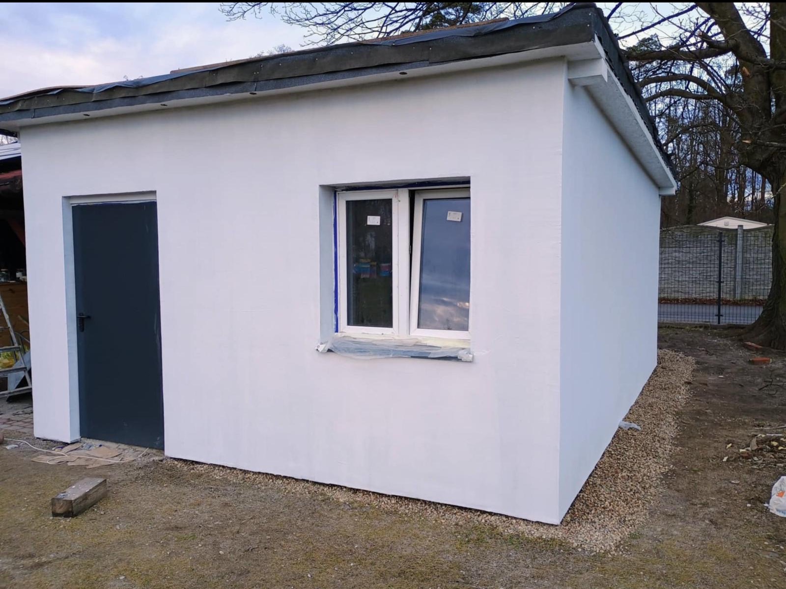Świeżo otynkowany i pomalowany na biało budynek gospodarczy z szarą stolarką drzwiową i oknem zabezpieczonym folią malarską, otoczony żwirem i ziemią.