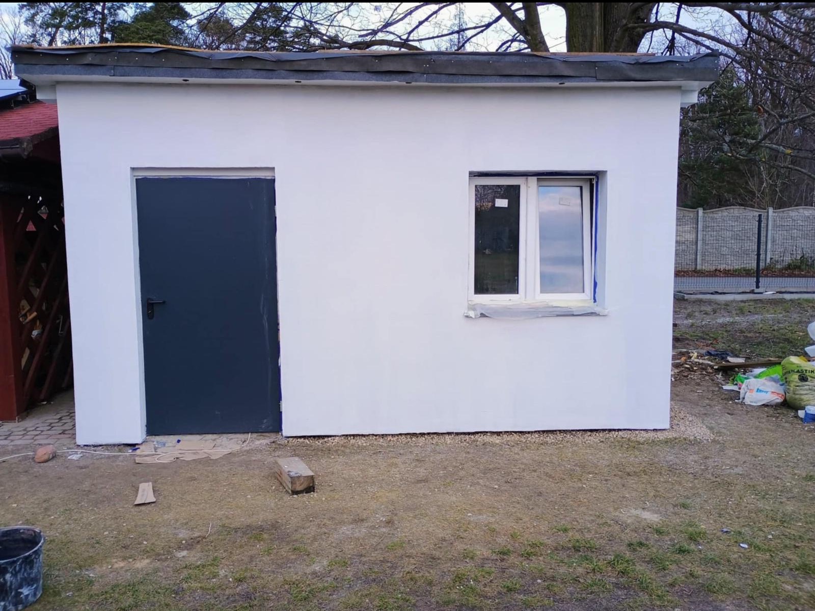Świeżo pomalowany na biało budynek gospodarczy z ciemnymi drzwiami i oknem, zabezpieczonym taśmą malarską, widoczne ślady prac wykończeniowych na zewnątrz.