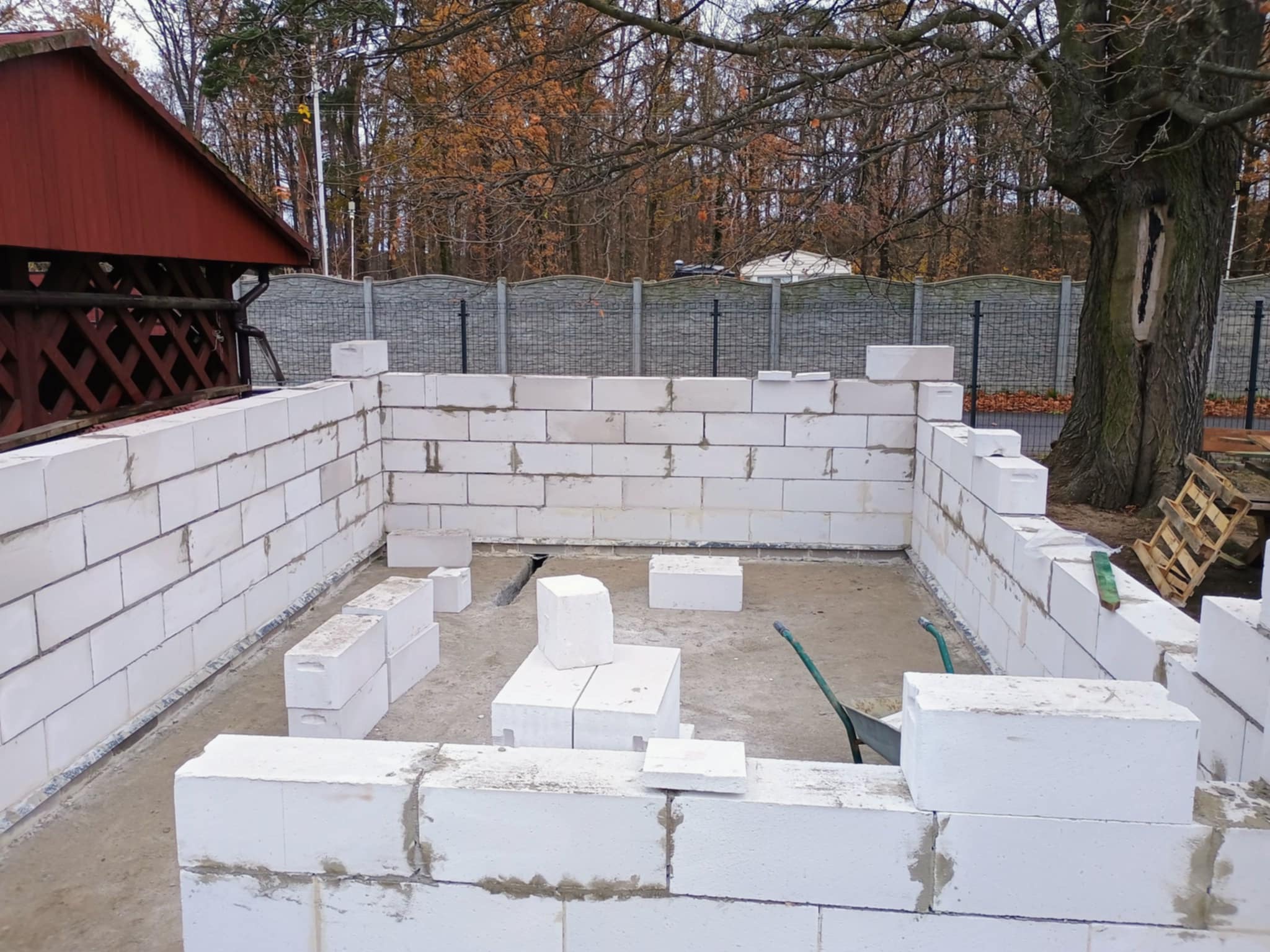 Wznoszenie ścian fundamentowych z białych bloczków betonowych na zewnątrz budynku; widoczna zaprawa, taczka i paleta w tle, drzewa bez liści.