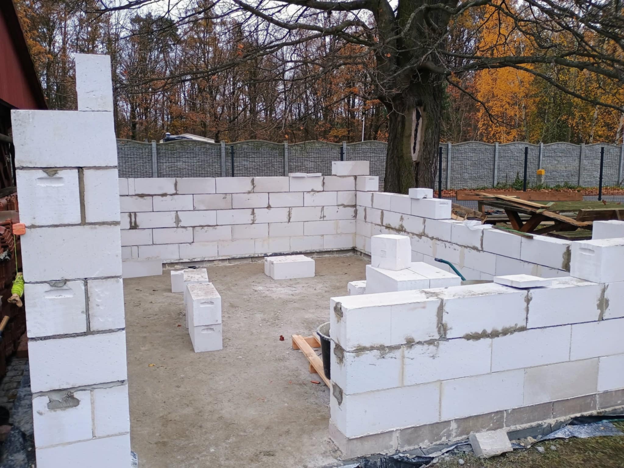 Wznoszenie ścian parterowego budynku z białych bloczków betonowych, widoczne spoiny, w tle drzewa i ogrodzenie z siatki.