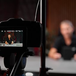 aim-Studio Sp. z o.o. - Ekran kamery z widokiem na nagrywaną osobę w studio. Widoczne ustawienia kamery i oznaczenie 'ON AIR'. W tle rozmyta sylwetka operatora.