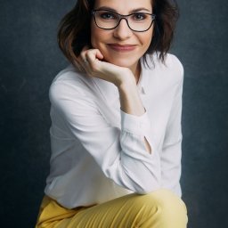 aim-Studio Sp. z o.o. - Portret kobiety w okularach, białej koszuli i żółtych spodni na ciemnym tle. Uśmiechnięta, z dłonią pod brodą, patrzy w obiektyw. Profesjonalna sesja.