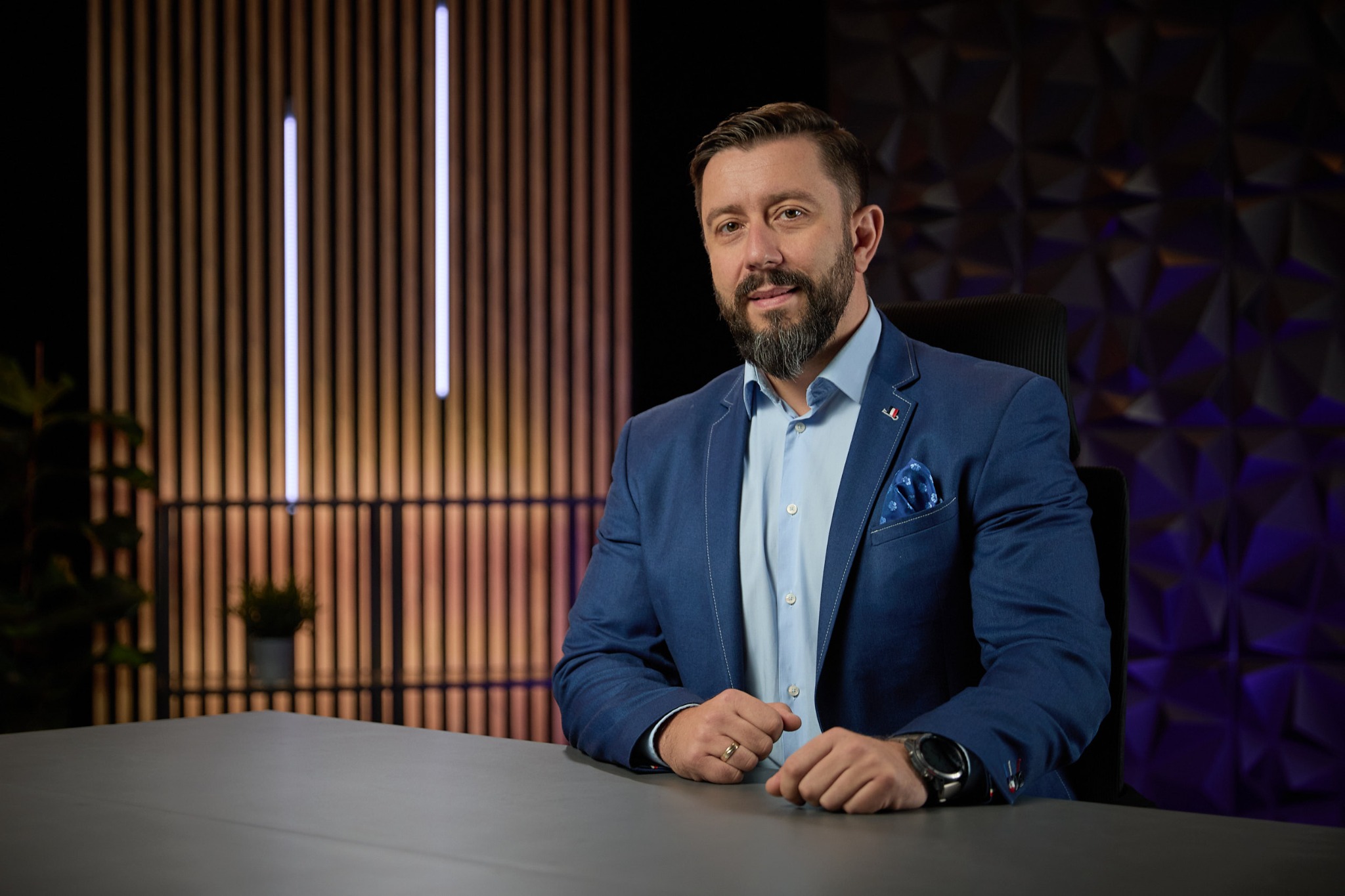 Portret mężczyzny w niebieskiej marynarce, siedzącego za biurkiem na tle dekoracyjnej ściany z podświetleniem LED i geometryczną strukturą 3D.