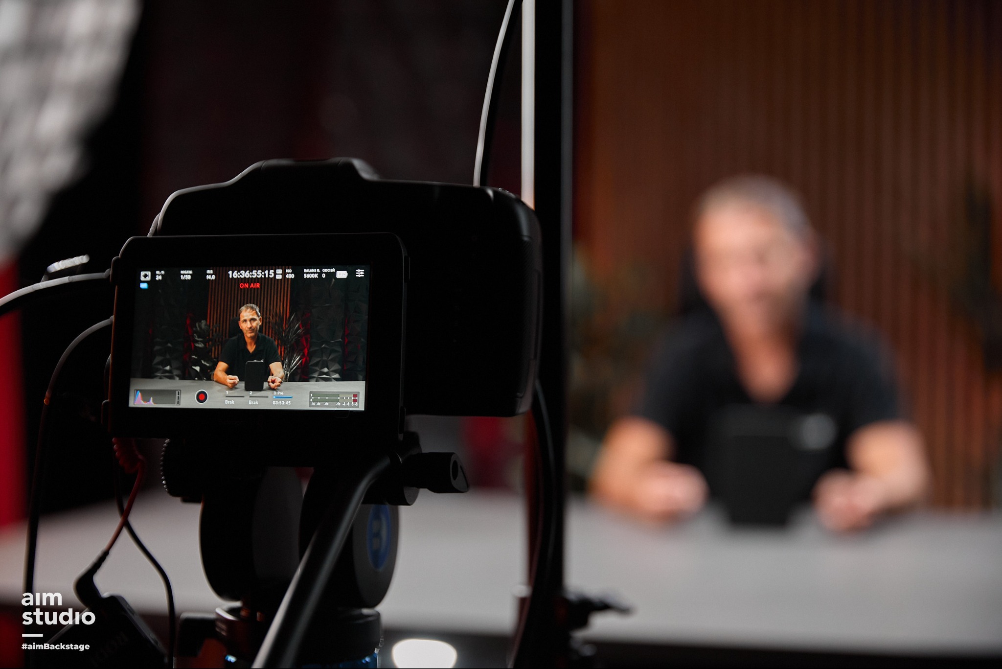 Ekran kamery z widokiem na nagrywaną osobę w studio. Widoczne ustawienia kamery i oznaczenie 'ON AIR'. W tle rozmyta sylwetka operatora.