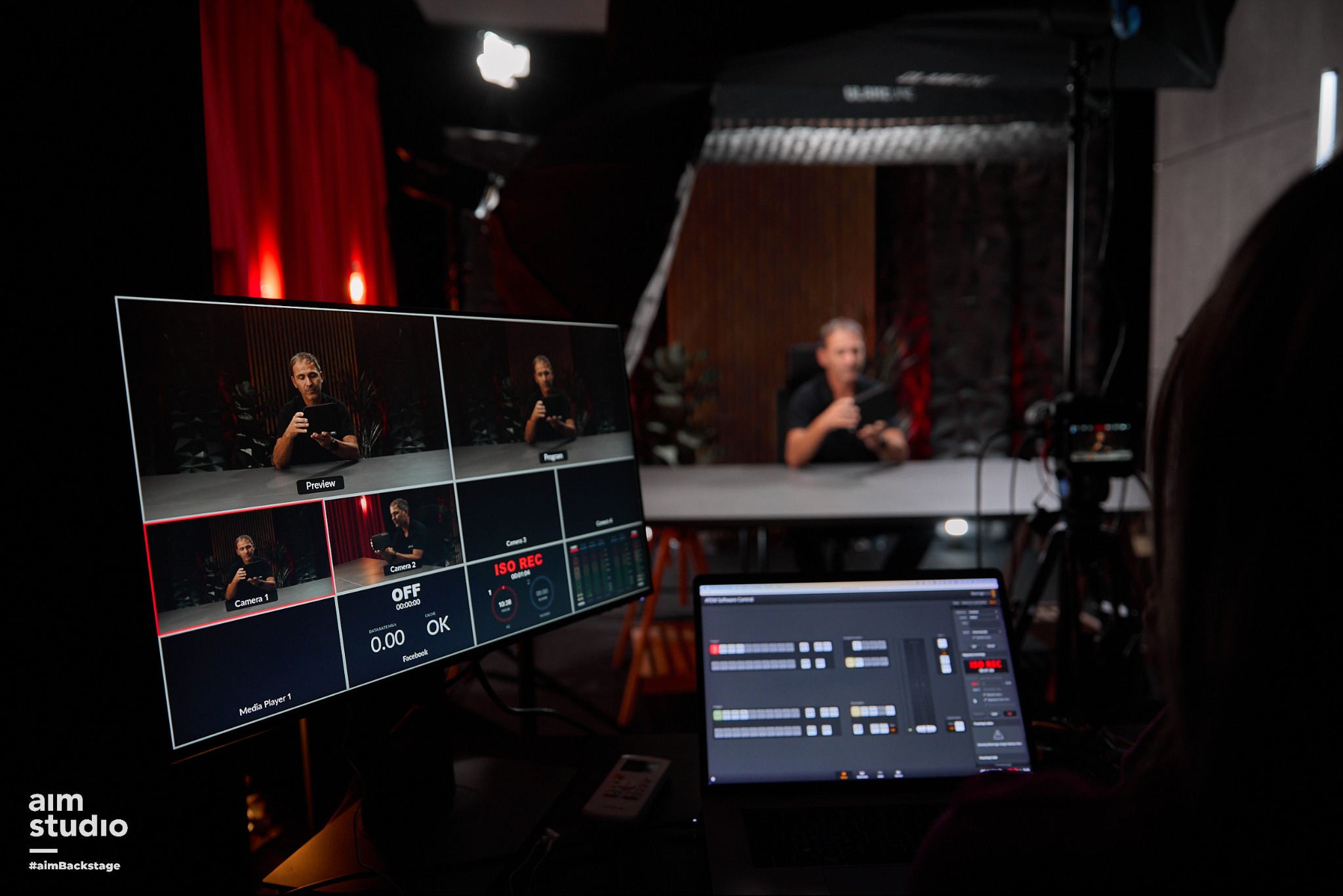 Studio filmowe: monitor z podglądem z kamer, laptop z oprogramowaniem do montażu, oświetlenie studyjne i osoba w tle, tworząca klimat profesjonalnej produkcji wideo.