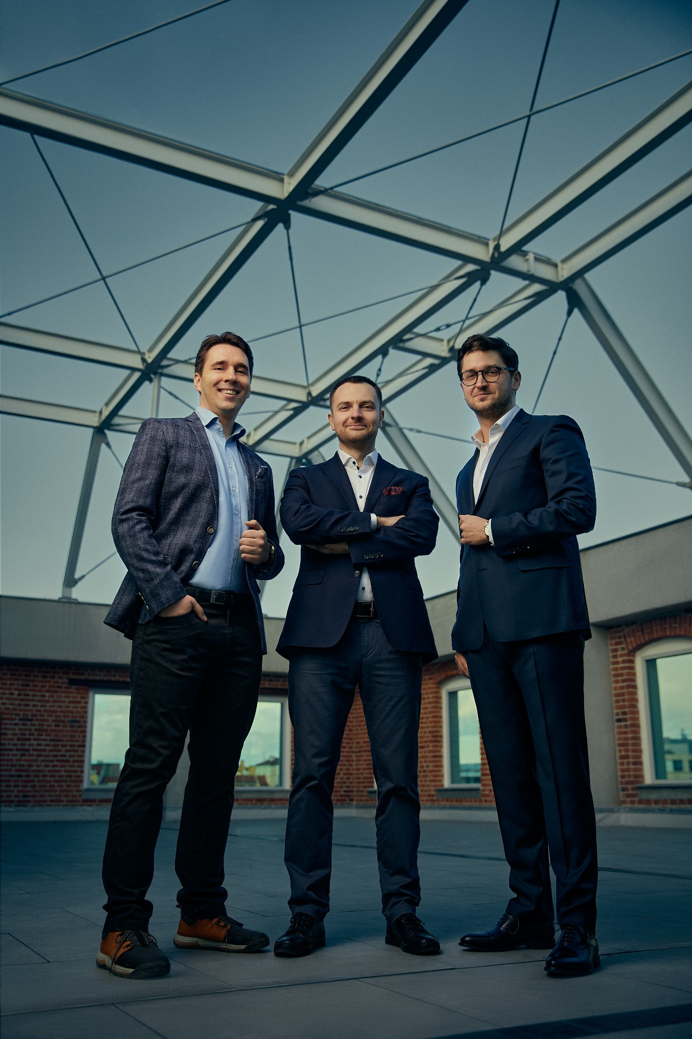 Trzech eleganckich mężczyzn w garniturach pozuje na tle nowoczesnej konstrukcji dachowej i ceglanego budynku. Uśmiechnięte twarze, pewne pozy. Profesjonalizm i nowoczesność.
