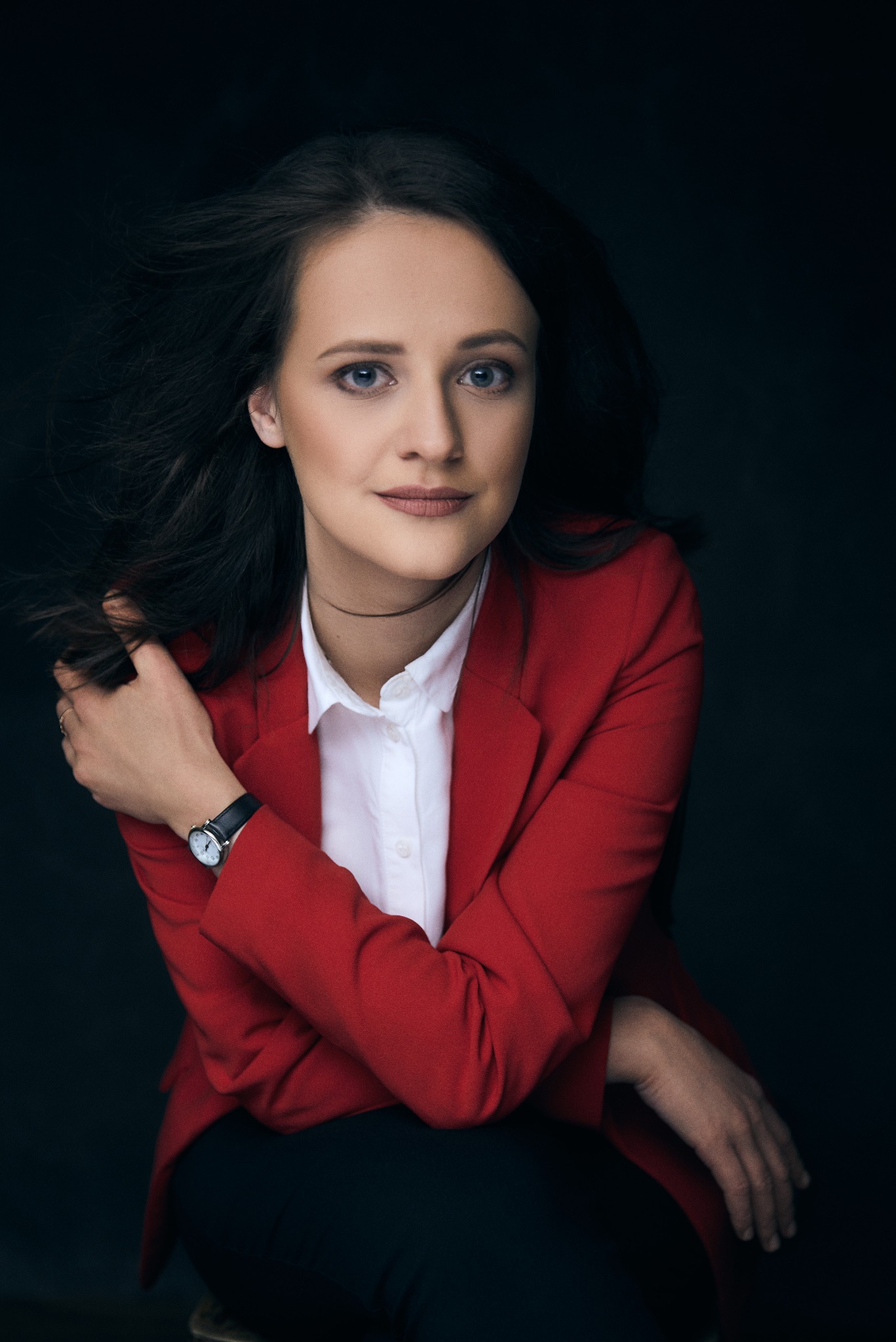 Portret kobiety w czerwonej marynarce na ciemnym tle, z delikatnym uśmiechem i włosami w ruchu, prezentujący profesjonalny wizerunek.