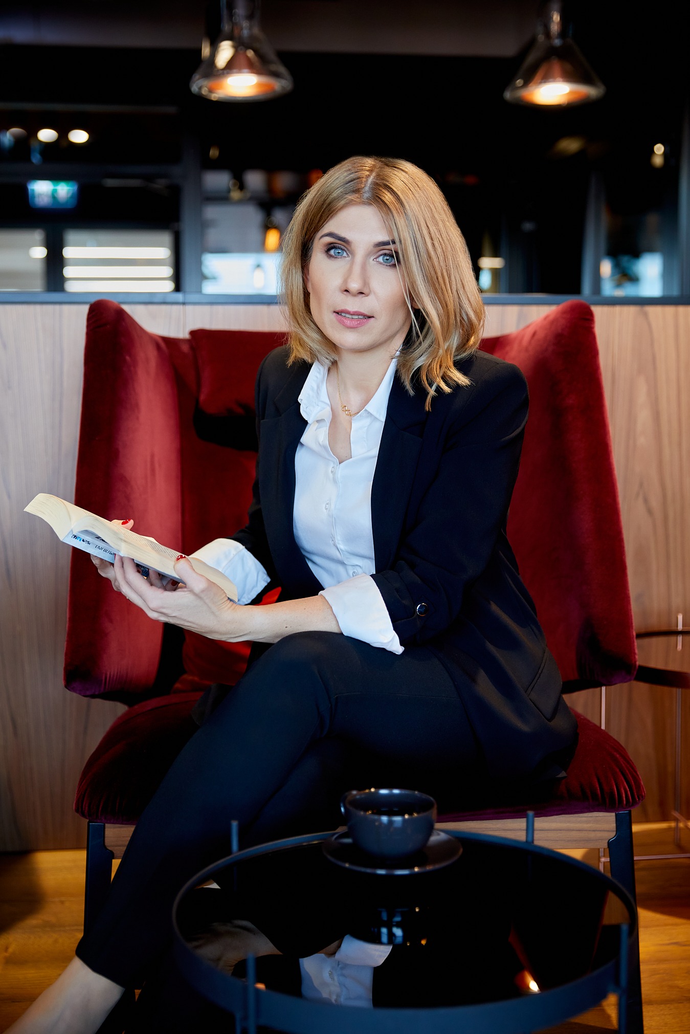 Portret kobiety w czarnym garniturze czytającej książkę w czerwonym fotelu, w kawiarni we Wrocławiu. Delikatne oświetlenie, spokojna atmosfera.